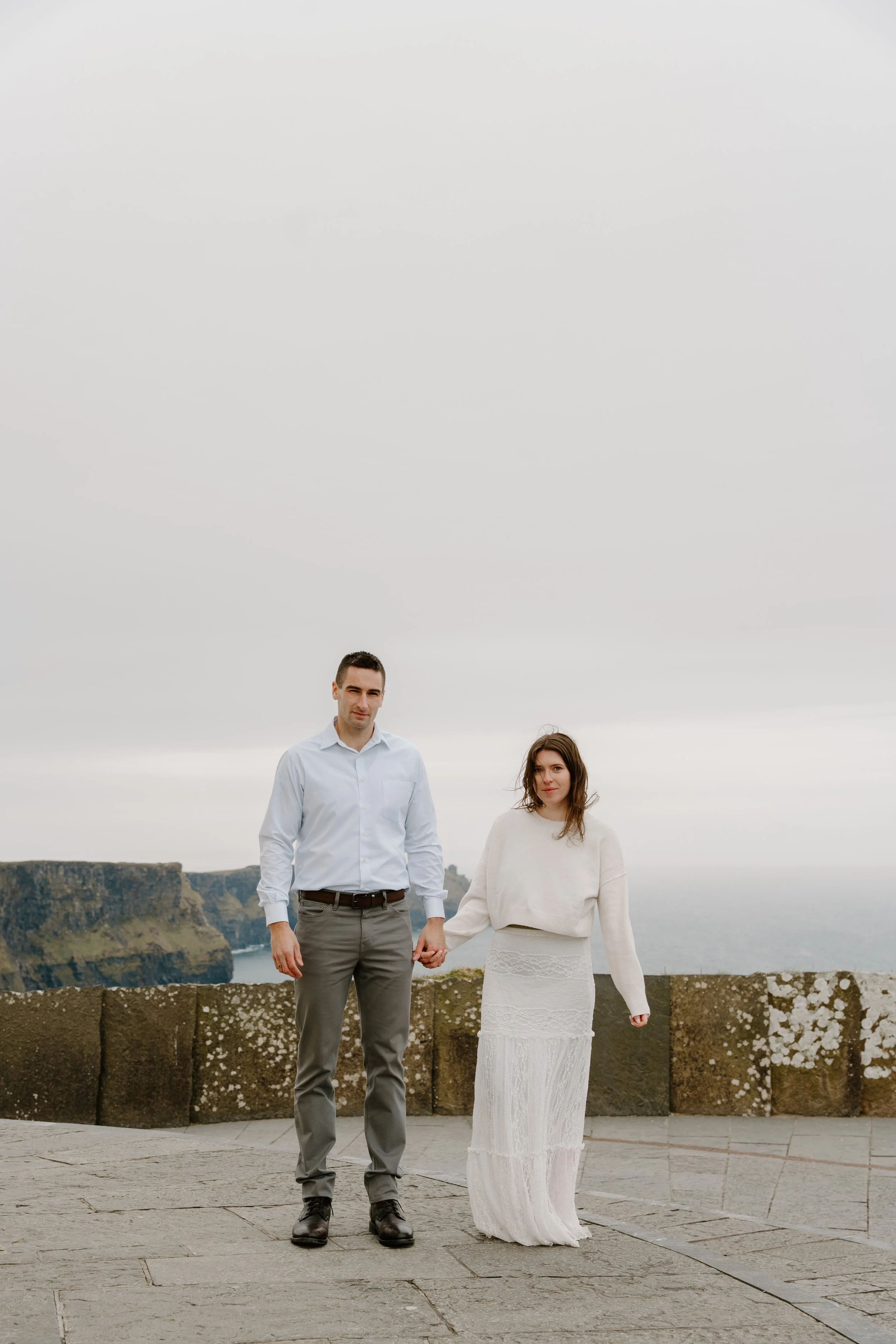 A couple standing on a stone-paved area holding hands with a cliff and cloudy sky in the background for an engagement photo session in Galway, Ireland. 
