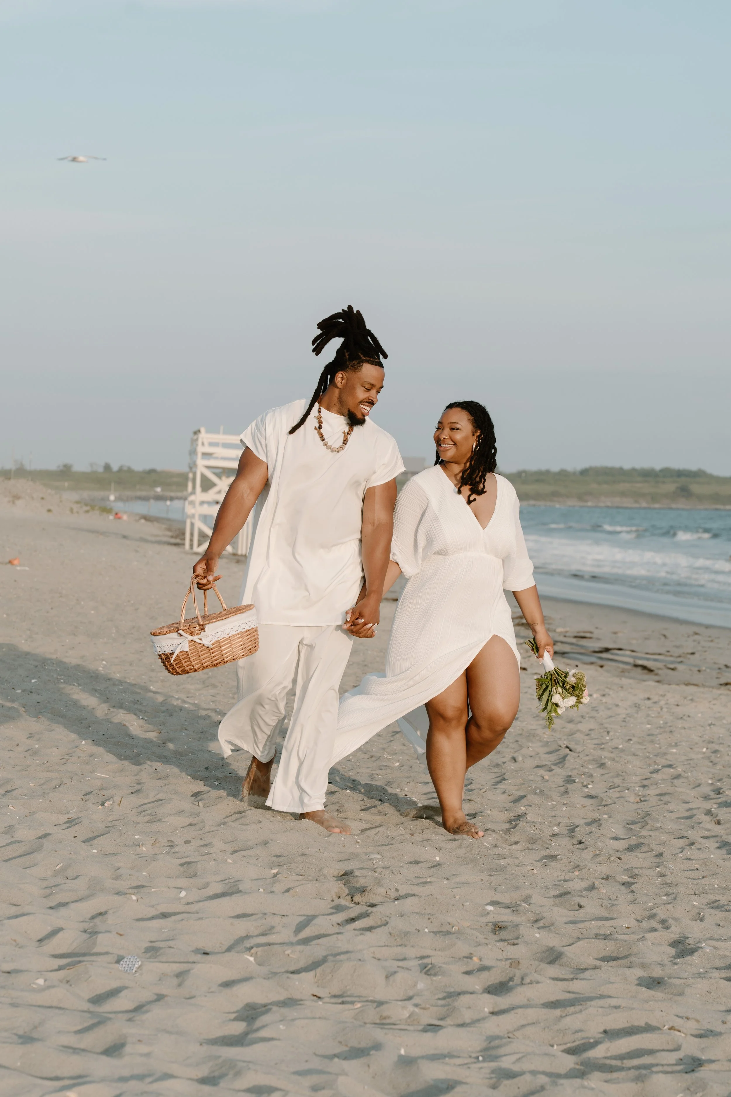 A smiling couple walking hand in hand along a beach, dressed in white, holding flowers and a picnic basket, with the ocean and sky in the background at an elopement at Second Beach in Rhode Island. 