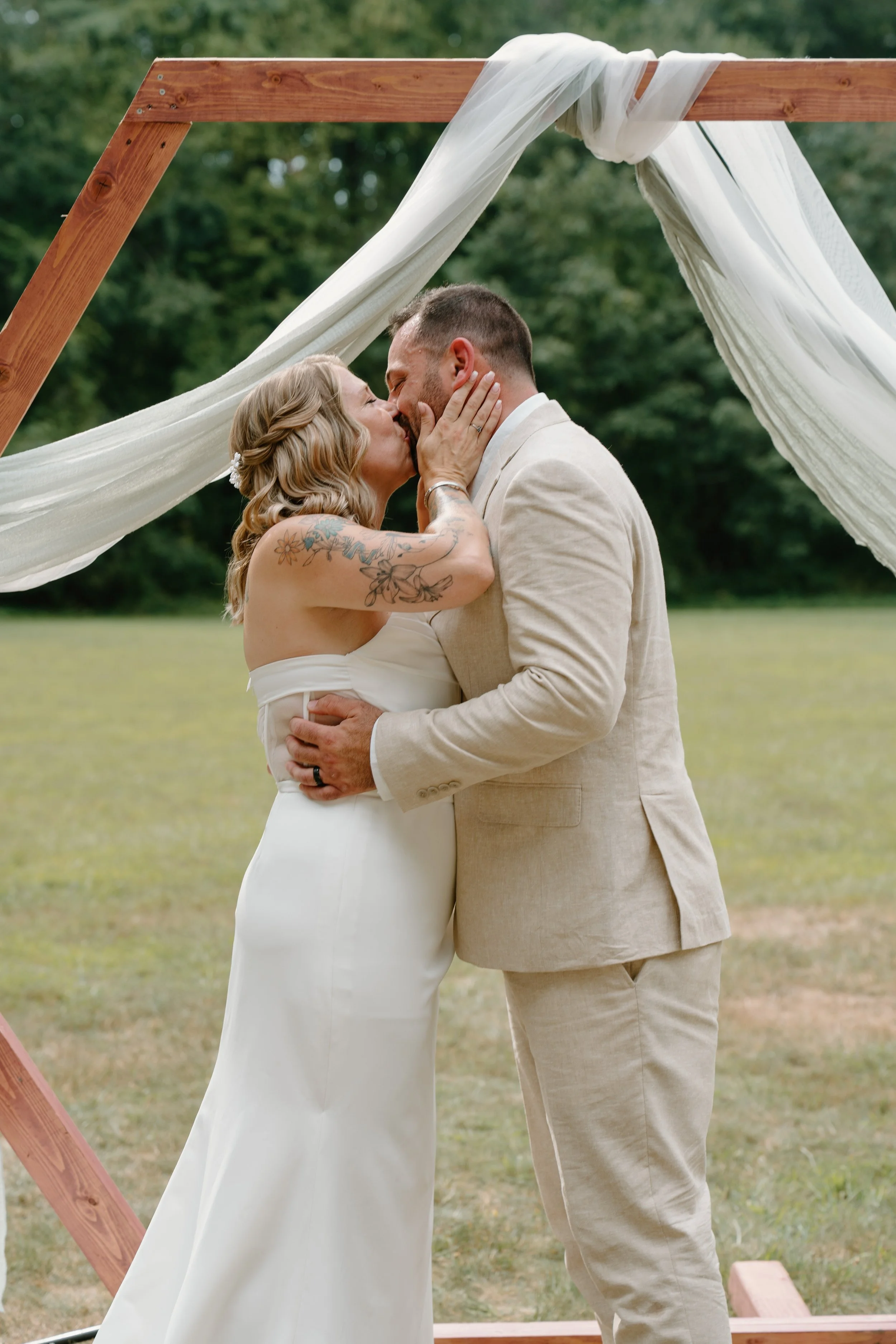A bride and groom kissing outdoors during their wedding ceremony beneath a wooden arch with white fabric drapes for a wedding at Tunxis Country Club in Connecticut. 