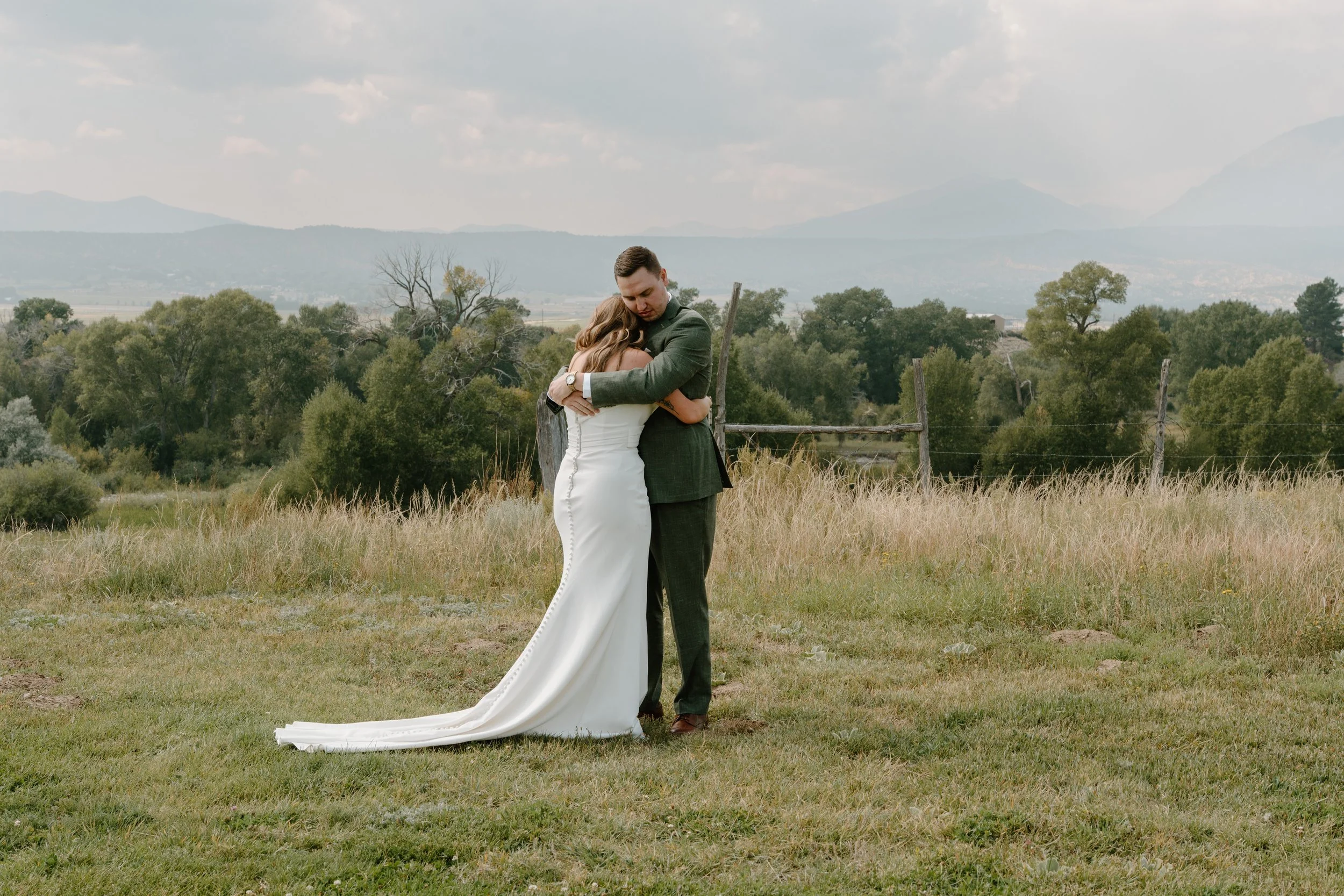 A bride and groom hugging outdoors in a grassy field with trees and mountains in the background at a wedding at Everett Ranch in Salida, Colorado.