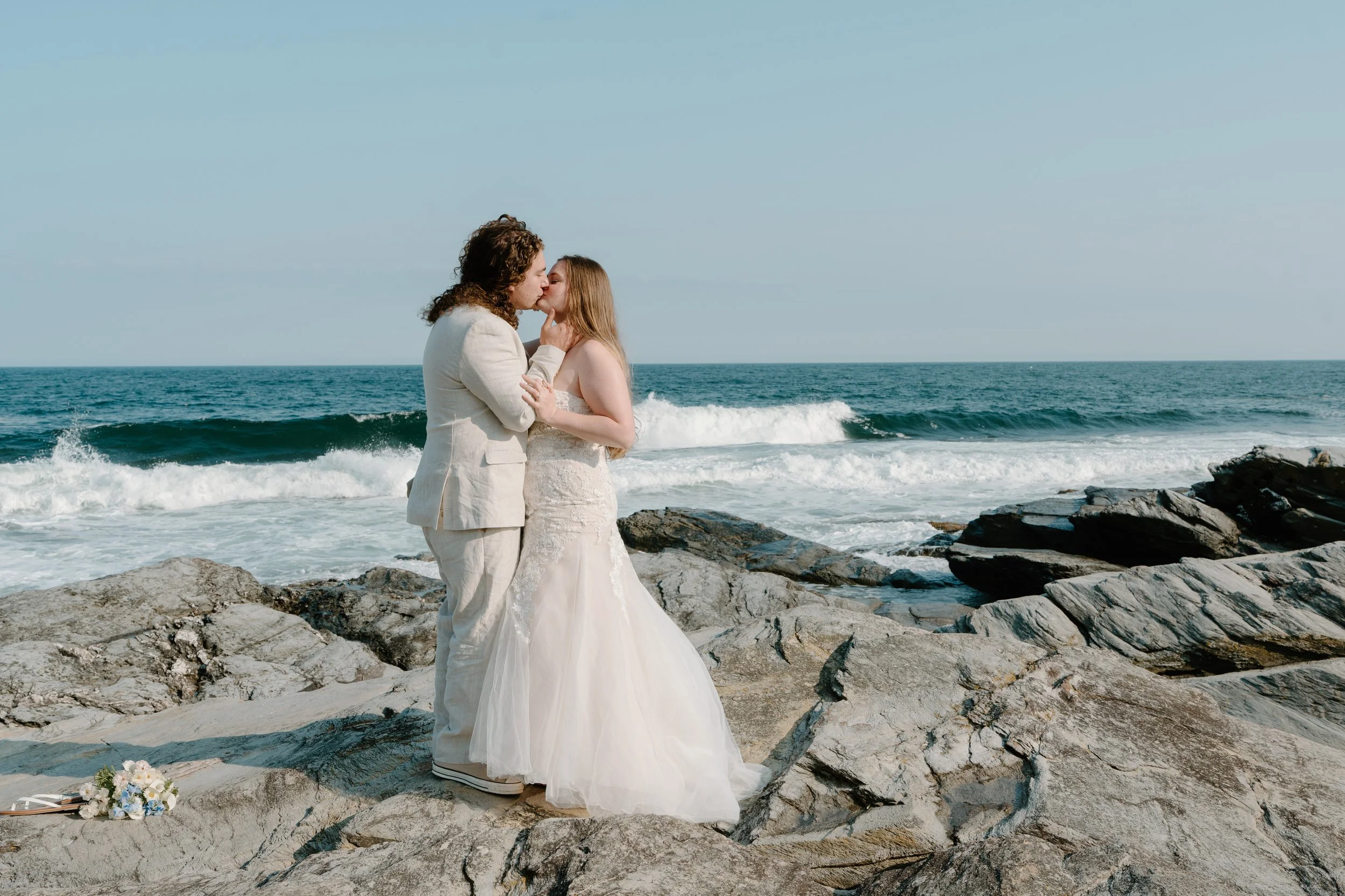A couple in wedding attire sharing a kiss on a rocky beach with waves and a clear sky in the background at an elopement at Beavertail State Park in Rhode Island. 