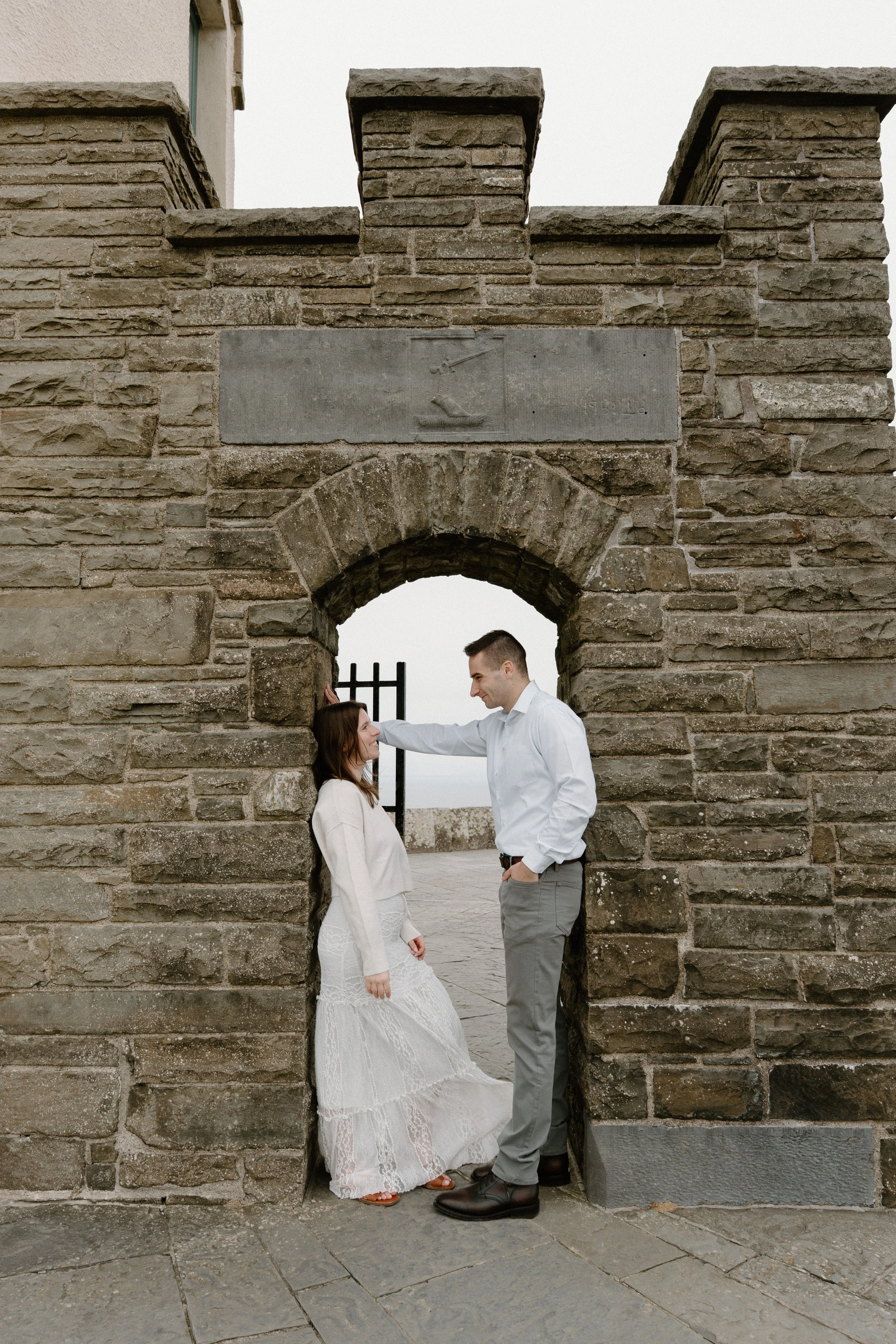 A couple standing in a stone archway on a cloudy day, with the woman in a white lace dress and the man in a white shirt and gray pants, sharing an intimate moment for an engagement photo session in Galway, Ireland. 