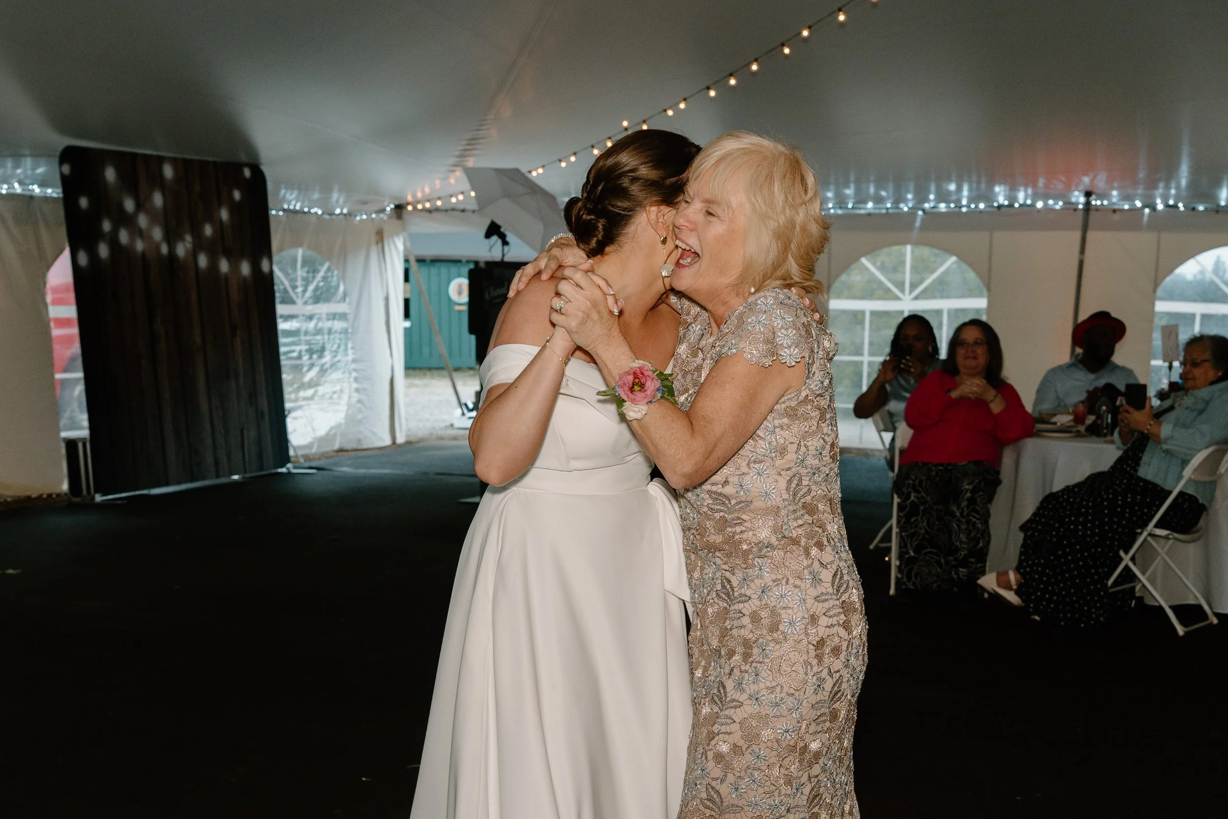 A bride in a white wedding dress and her mother in a floral dress share a joyful moment at a wedding reception, hugging closely and smiling, inside a tent with seated guests in the background for a wedding at Holiday Hill Day Camp in Connecticut. 