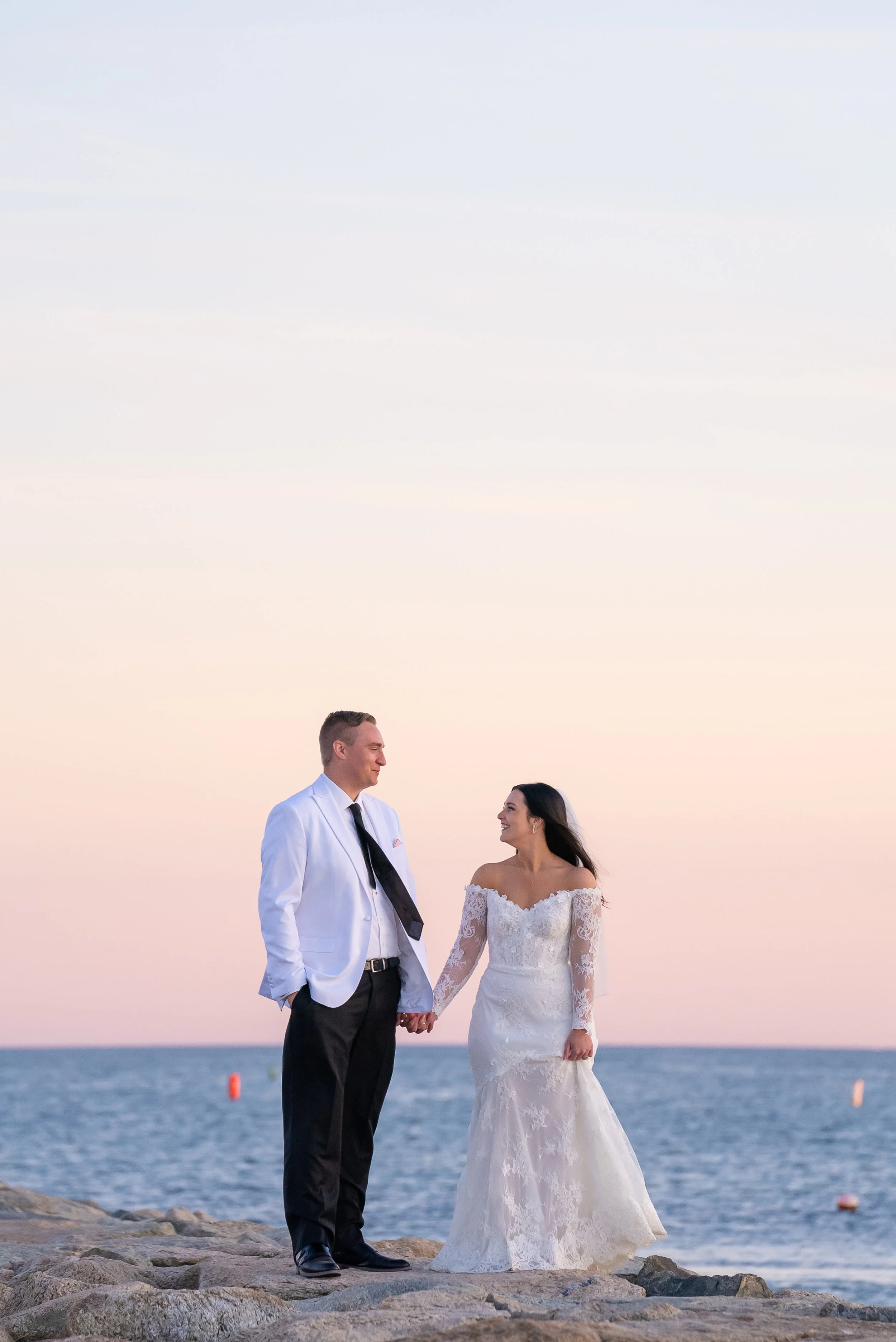 A bride and groom holding hands on rocks by the ocean during sunset, smiling at each other during a wedding at Red Jacket Resort in Cape Cod. 