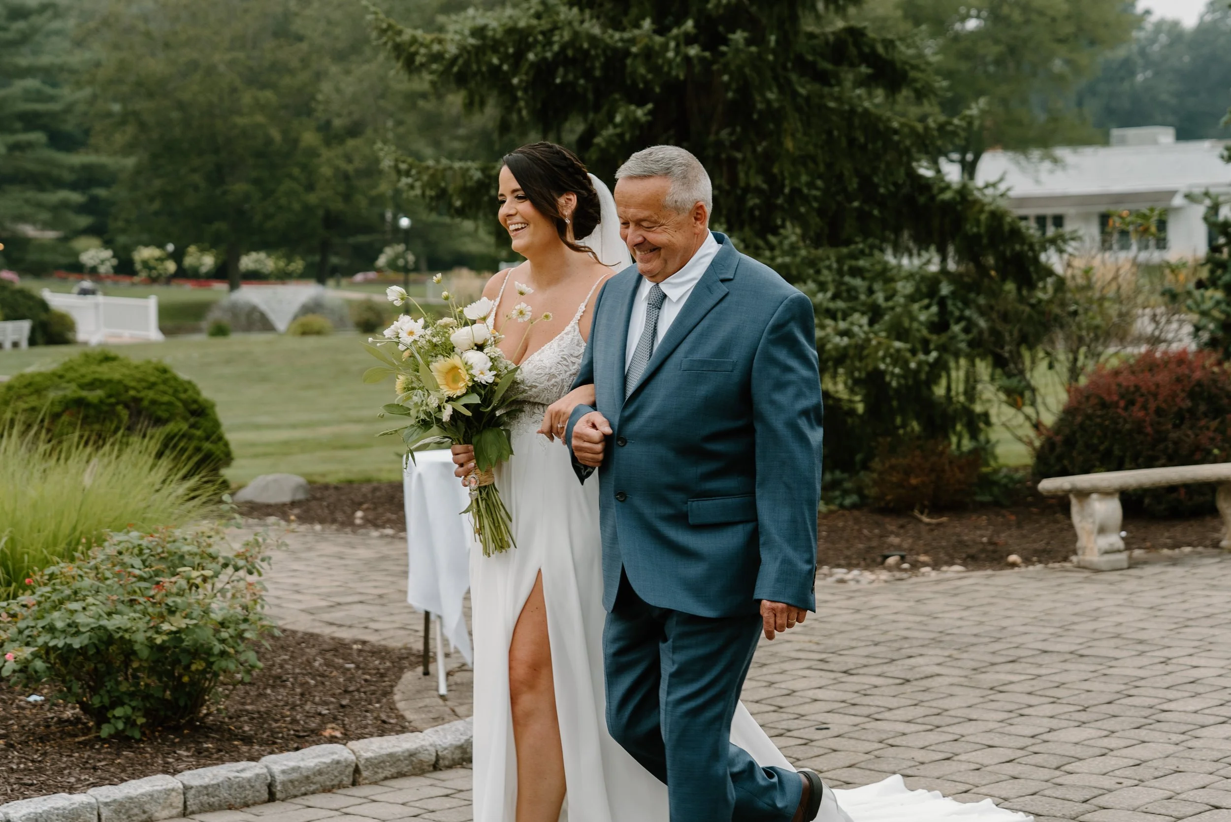 Bride walking down the aisle arm-in-arm with an older man in a suit, during an outdoor wedding ceremony, smiling with trees and a garden in the background during a wedding at the Aqua Turf Club in Connecticut. 