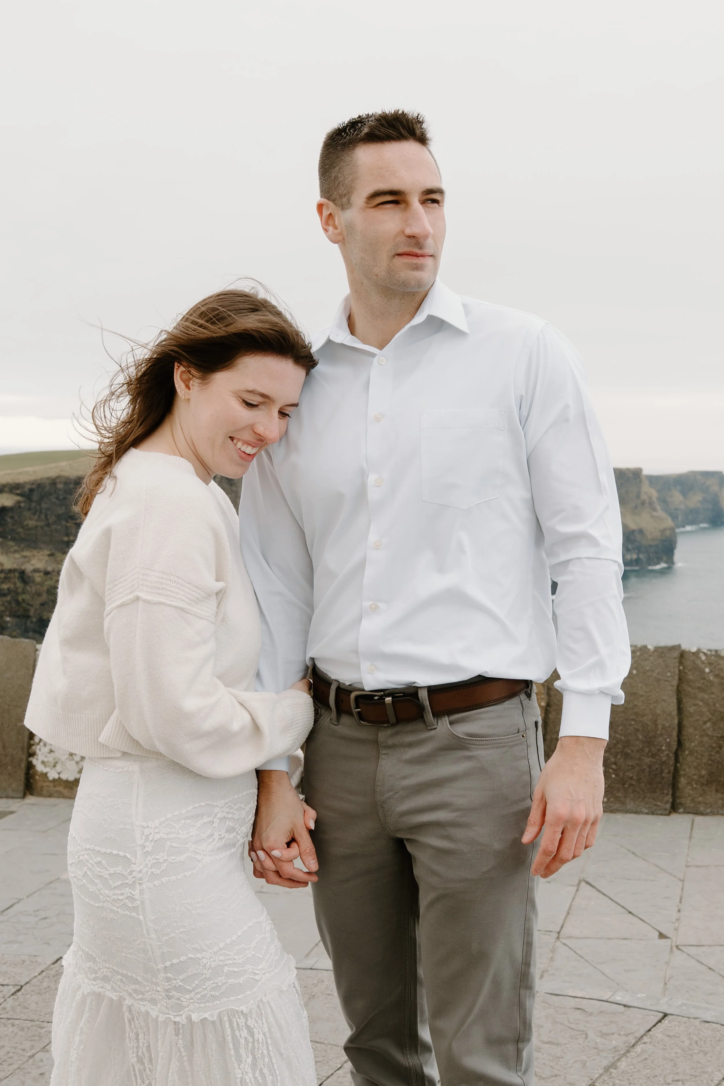 A man and woman holding hands, standing outdoors with cliffs and ocean in the background, dressed casually and smiling for an engagement photo session in Galway, Ireland. 