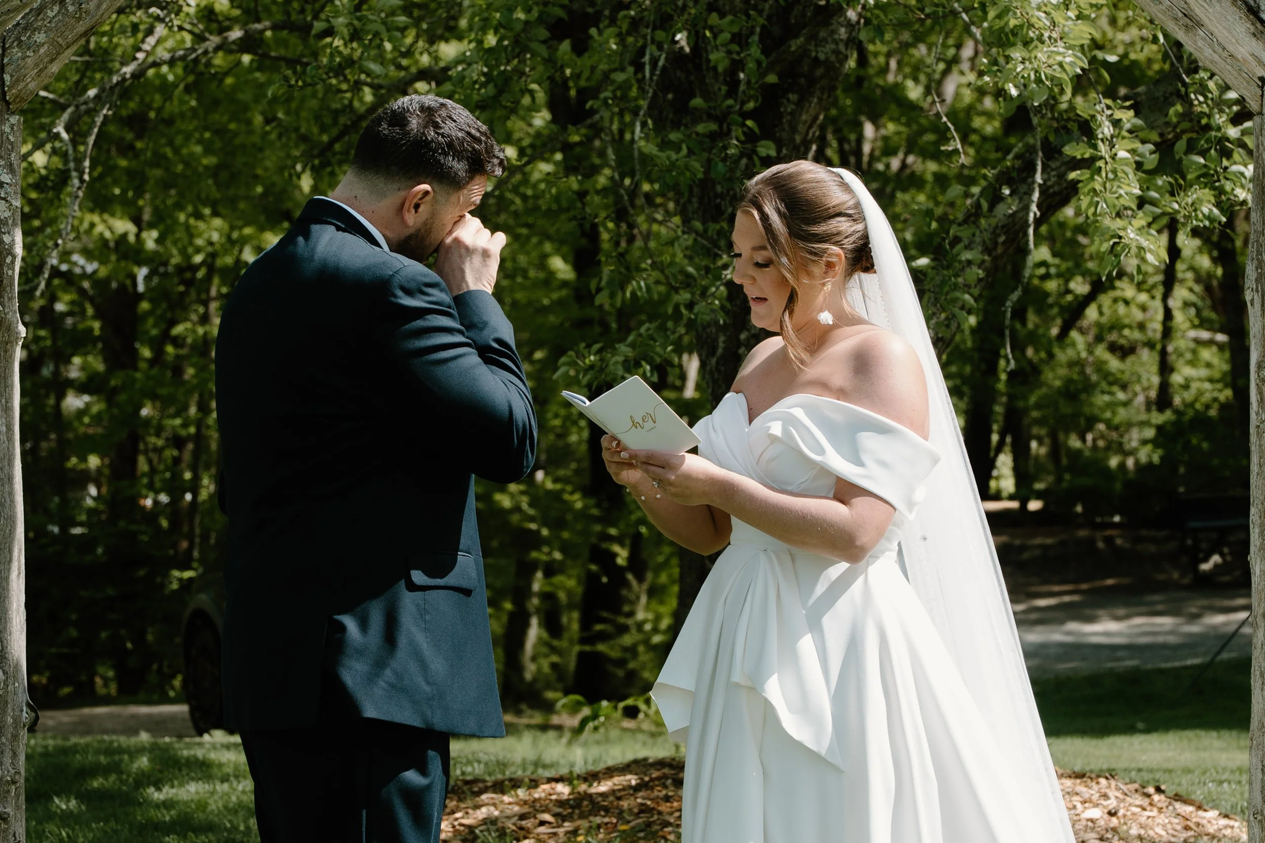 A bride and groom exchanging vows at an outdoor wedding ceremony surrounded by green trees, with the bride reading from a small book and the groom wiping his eyes for a wedding at Holiday Hill Day Camp in Connecticut.