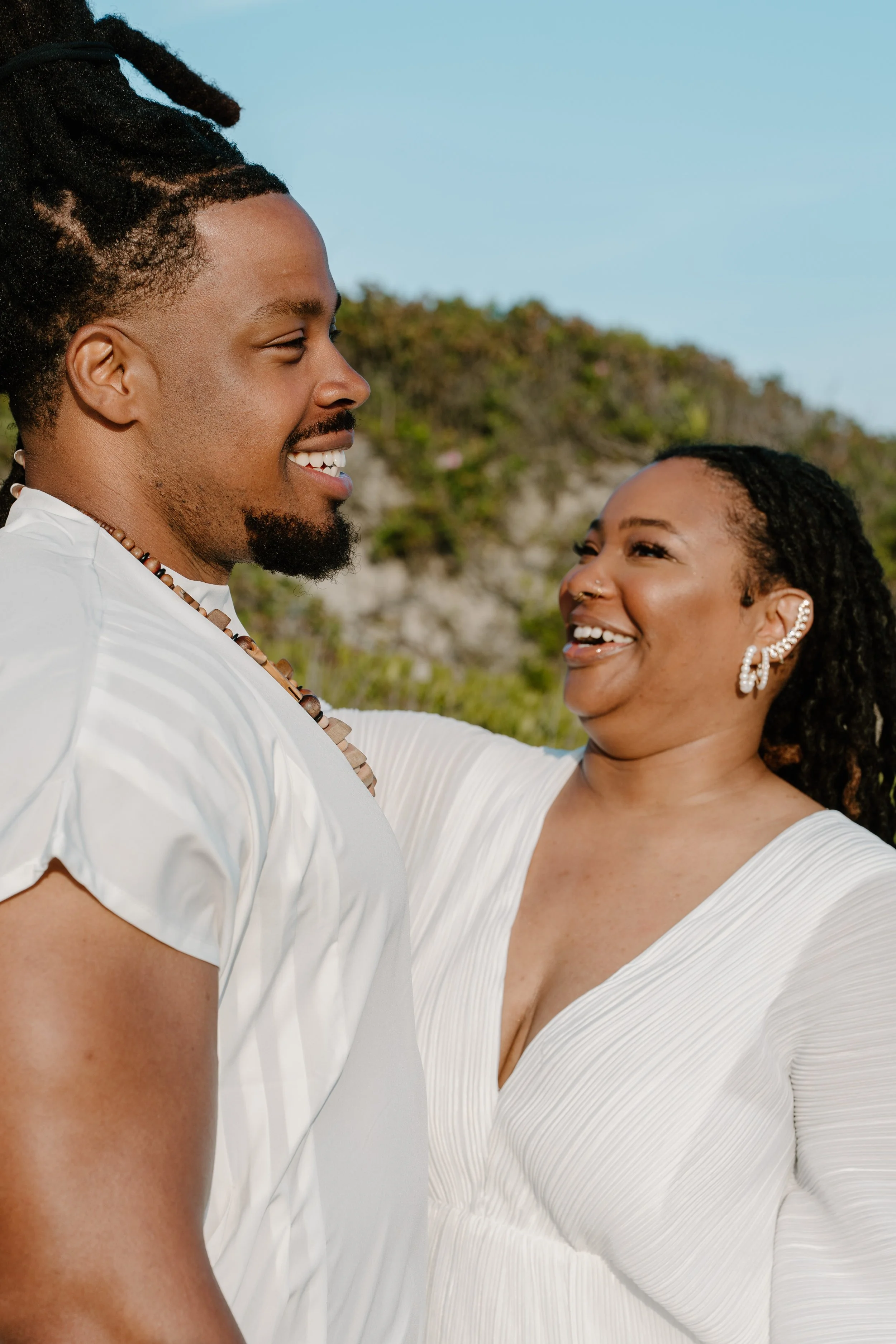 A couple smiling and looking at each other outdoors with a rocky hillside and blue sky in the background at an elopement at Second Beach in Rhode Island. 