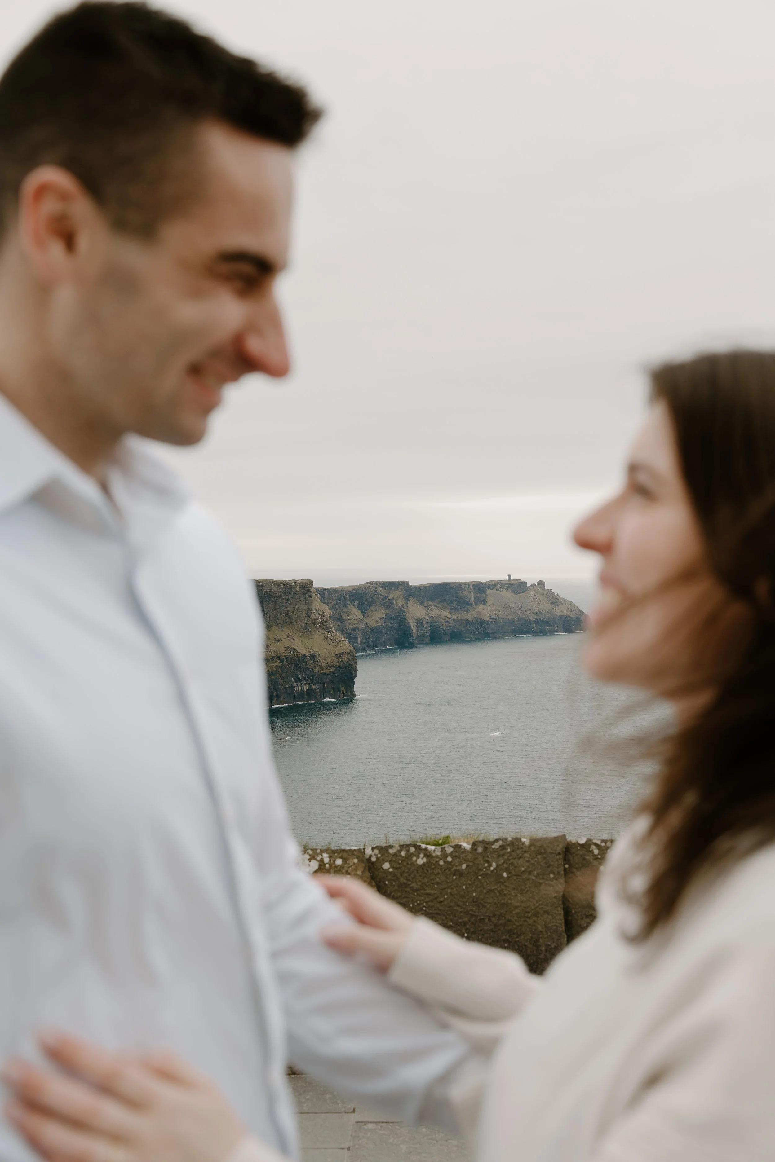 A man and woman standing close together with the ocean and cliffs in the background, smiling at each other for an engagement photo session in Galway, Ireland. 