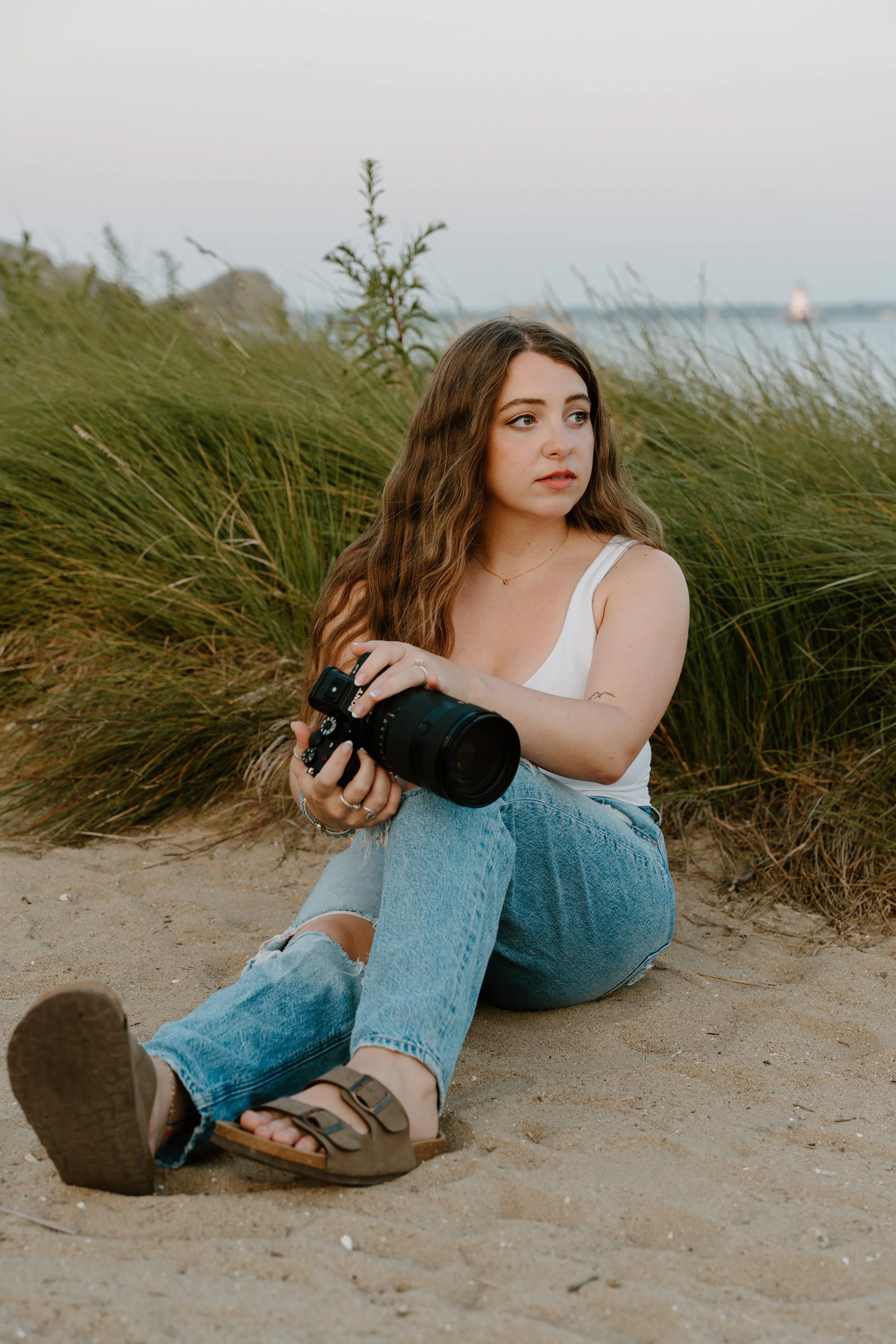 Kaylee of Unlost Media sits on a sandy beach, holding a camera.