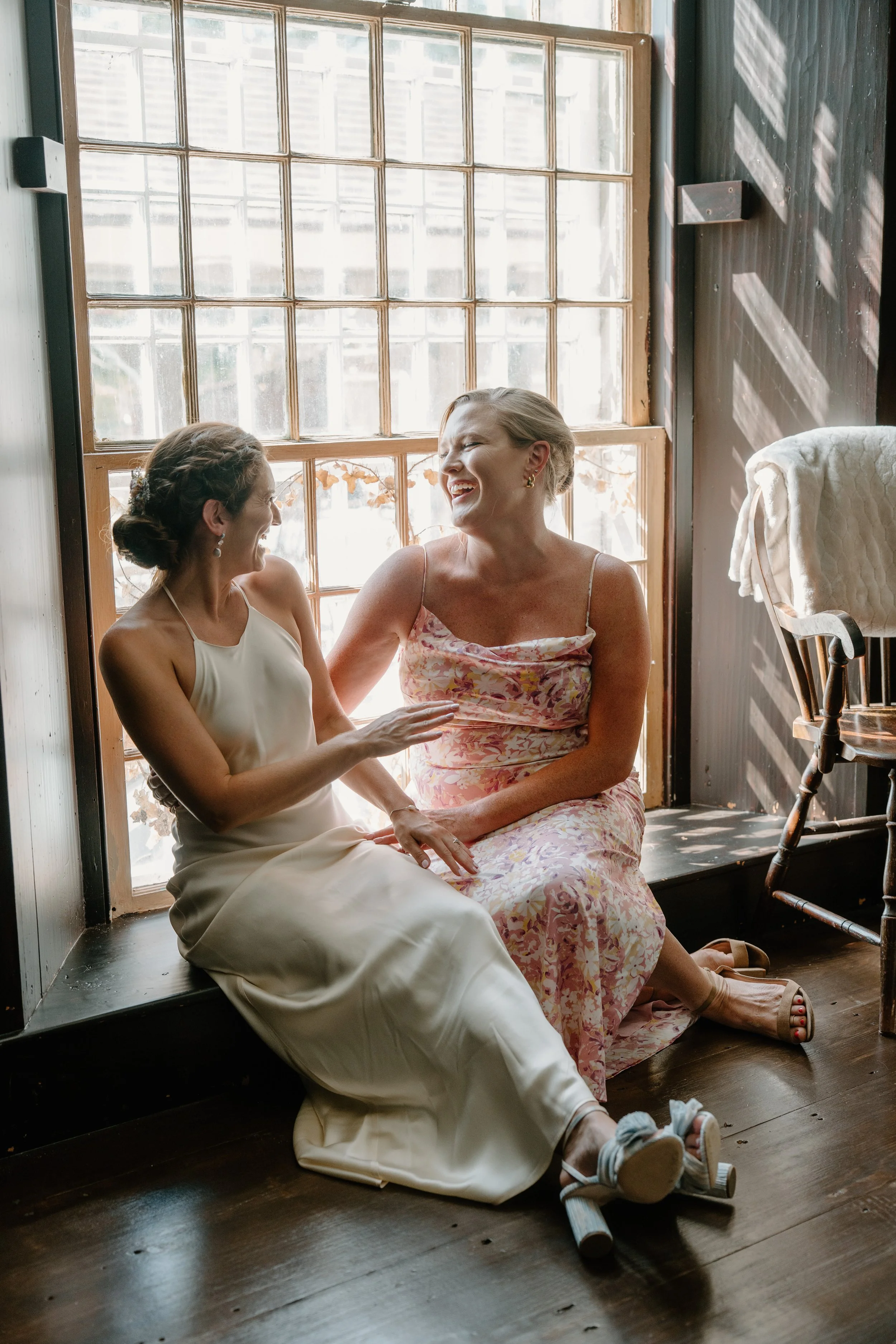 Two women sitting on a window seat, laughing and talking warmly, with sunlight streaming in through a large grid window, in a cozy room at a wedding in Bath, Maine. 