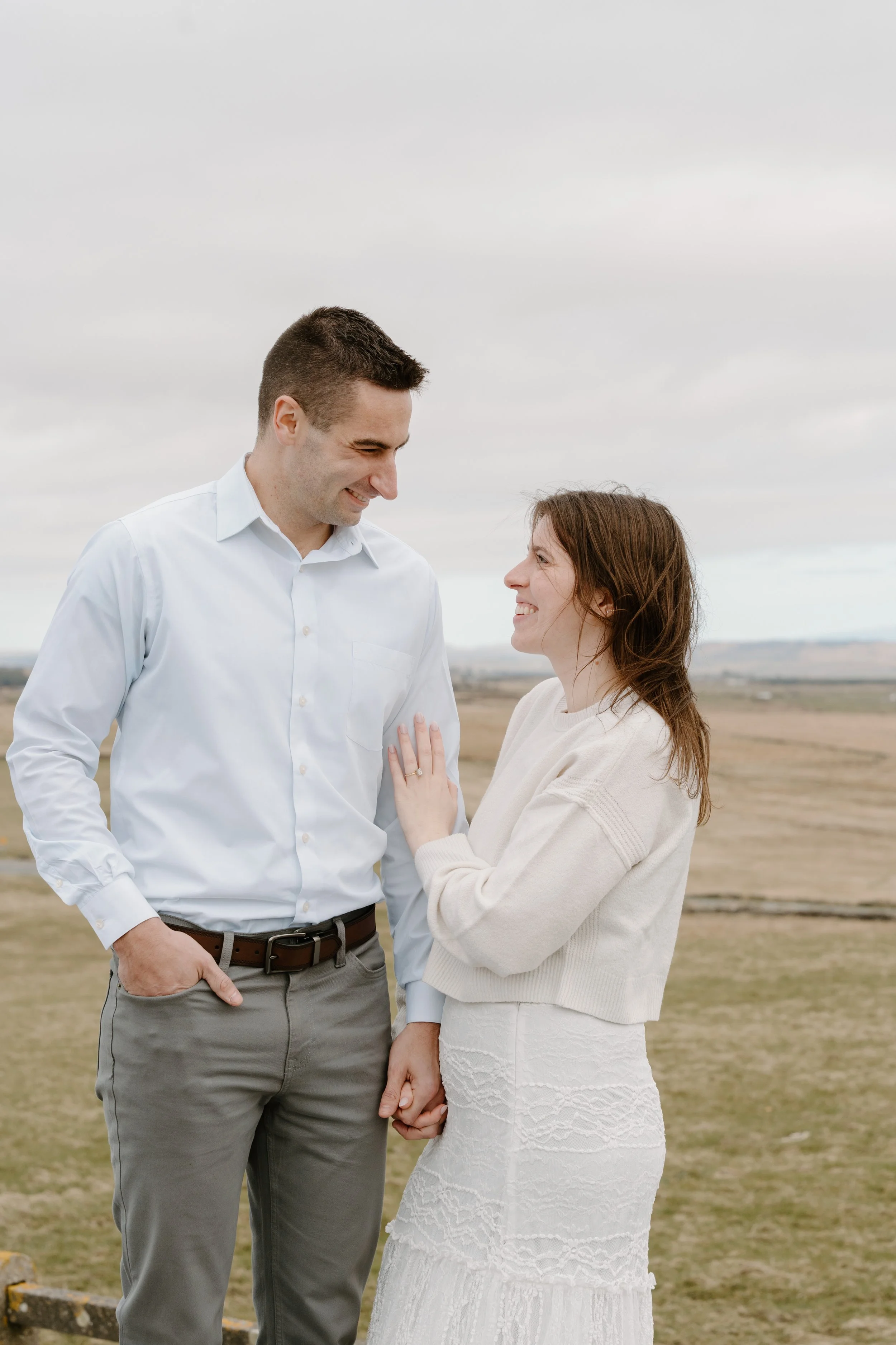 A couple smiling and holding hands outdoors on a cloudy day, facing each other for an engagement photo session in Galway, Ireland. 