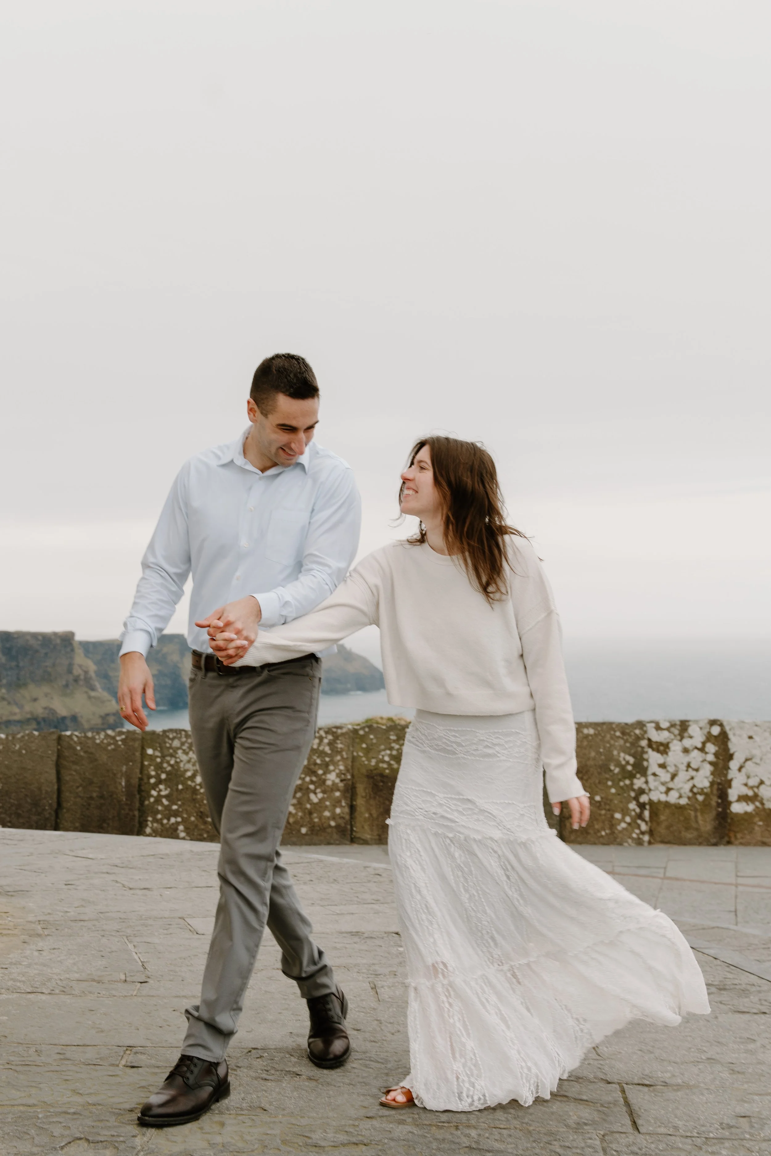 A couple dancing outdoors on a cloudy day, with cliffs and water in the background. They are smiling and holding hands, dressed in casual yet elegant clothing for an engagement photo session in Galway, Ireland. 