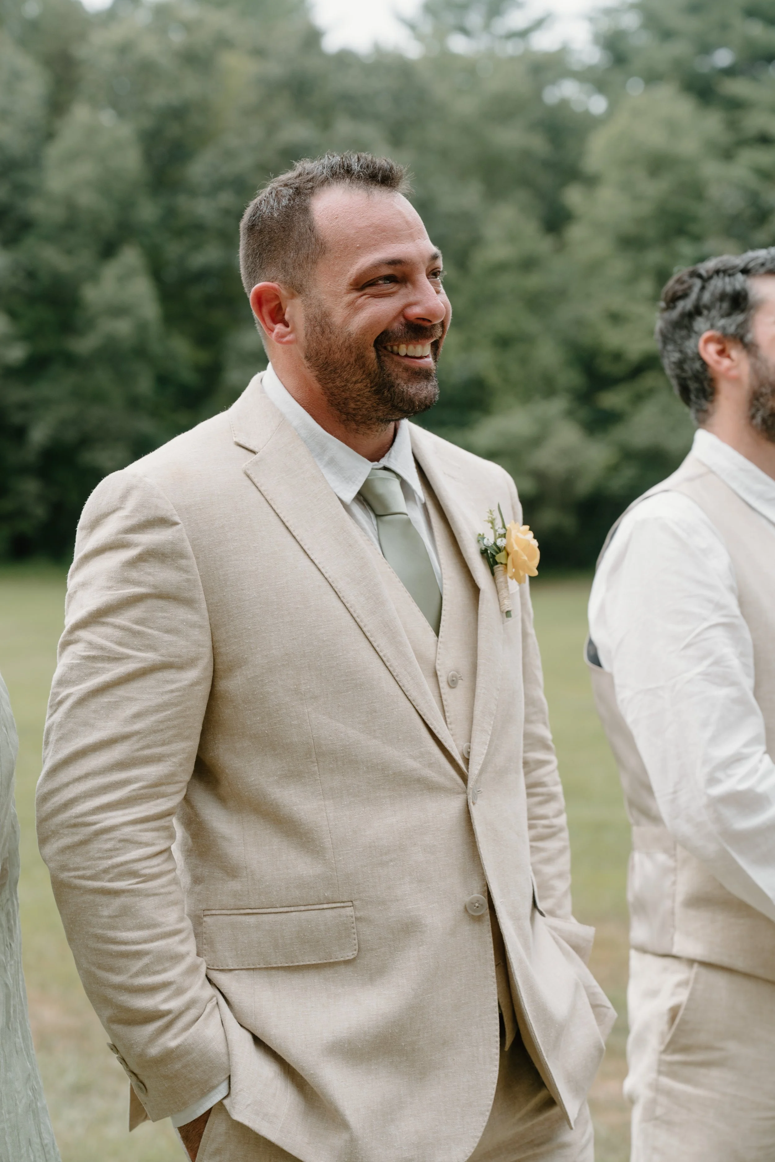 A man in a beige suit and tie smiling outdoors at a wedding or formal event, with other people in similar attire nearby and a background of trees for a wedding at Tunxis Country Club in Connecticut. 