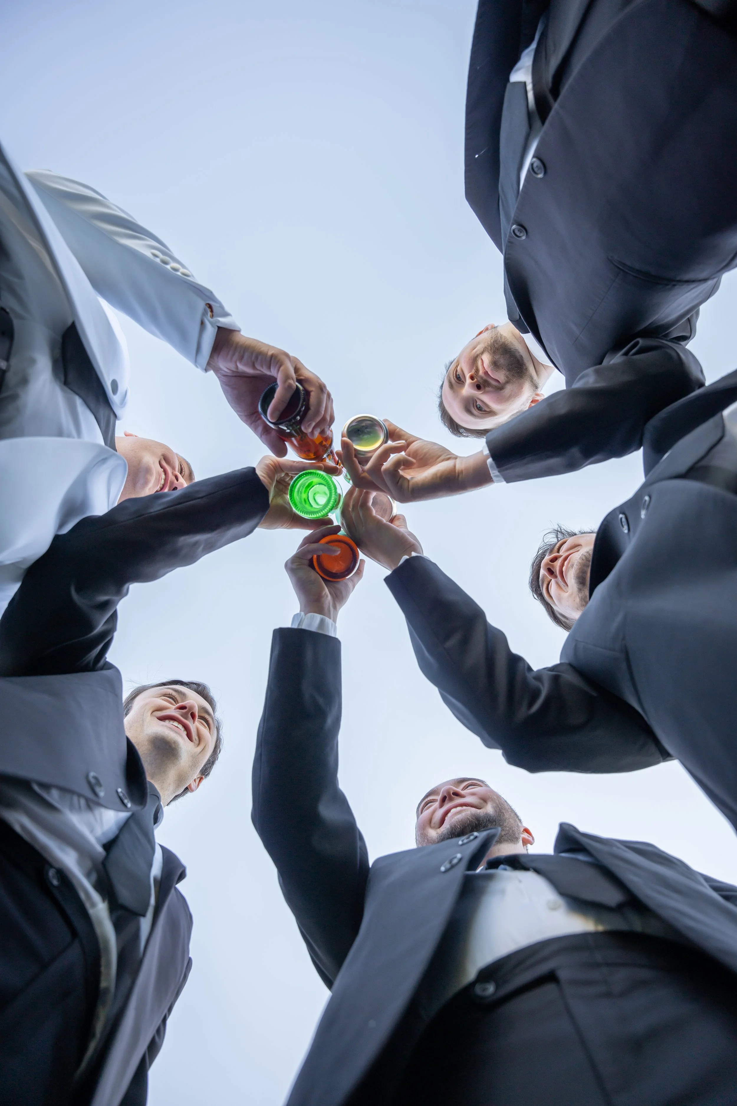 Group of men in suits and white coats holding colorful drinks in a circle, viewed from ground level against a cloudy sky during a wedding at Red Jacket Resort in Cape Cod. 