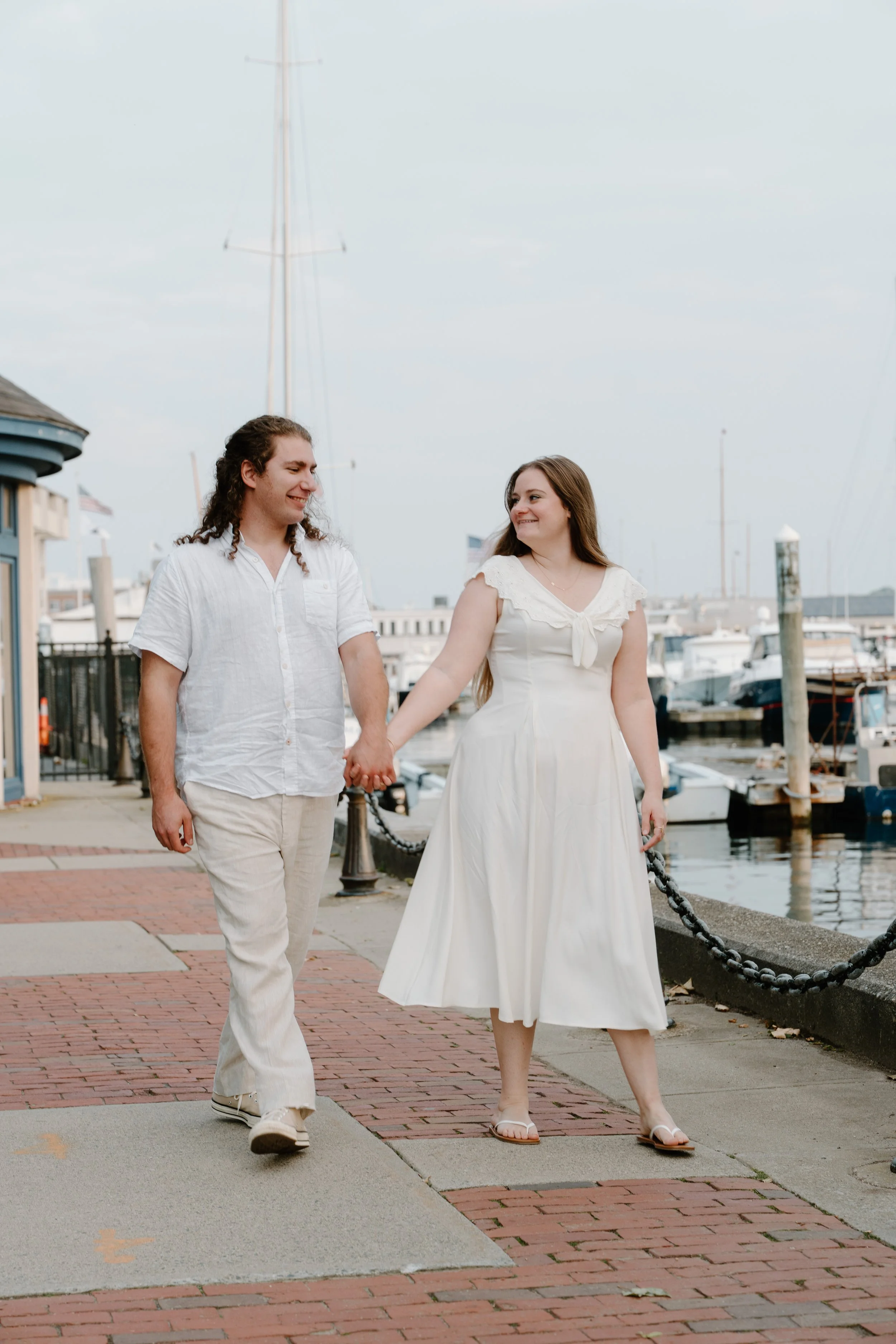 A happy couple walking hand in hand along a marina promenade, holding hands and smiling at each other at an elopement at Beavertail State Park in Rhode Island. 