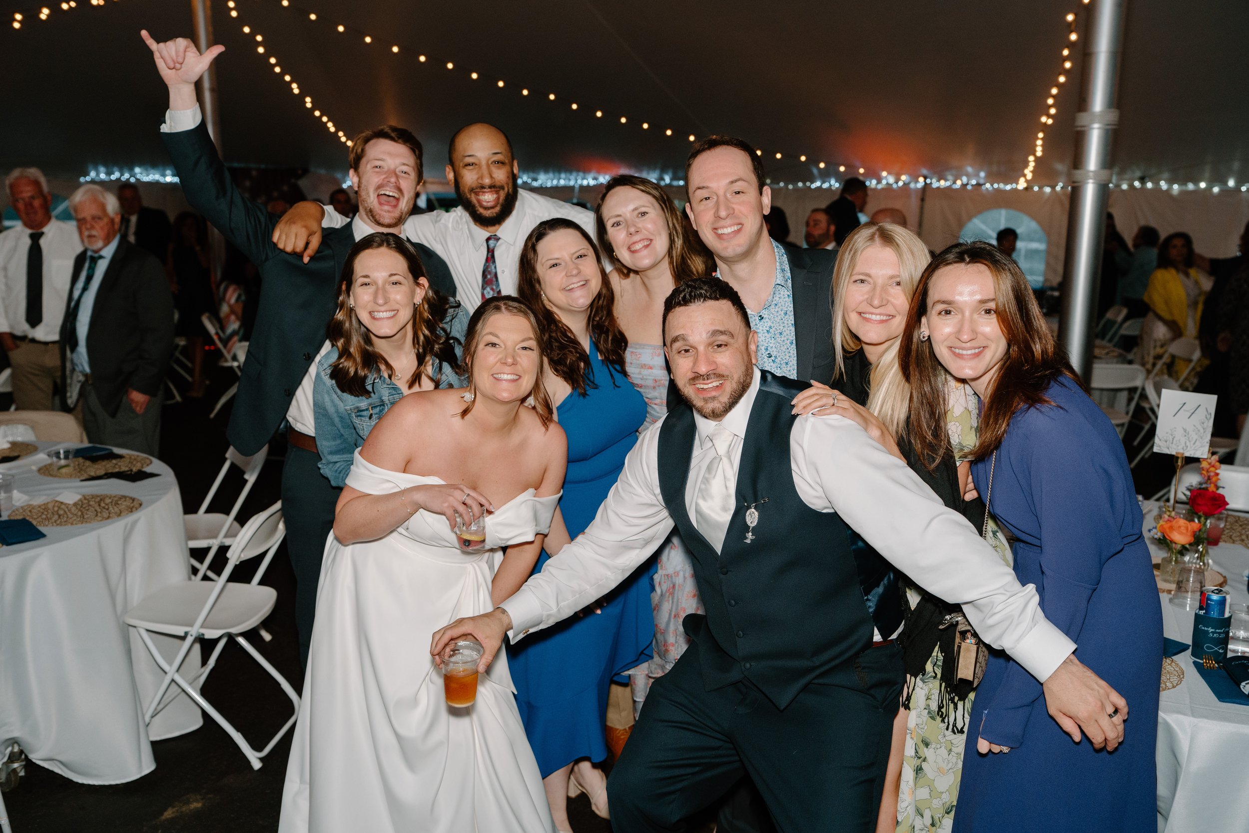 A group of people at a celebration or wedding in a tent, smiling and posing for the camera, with a decorated table and string lights in the background for a wedding at Holiday Hill Day Camp in Connecticut.