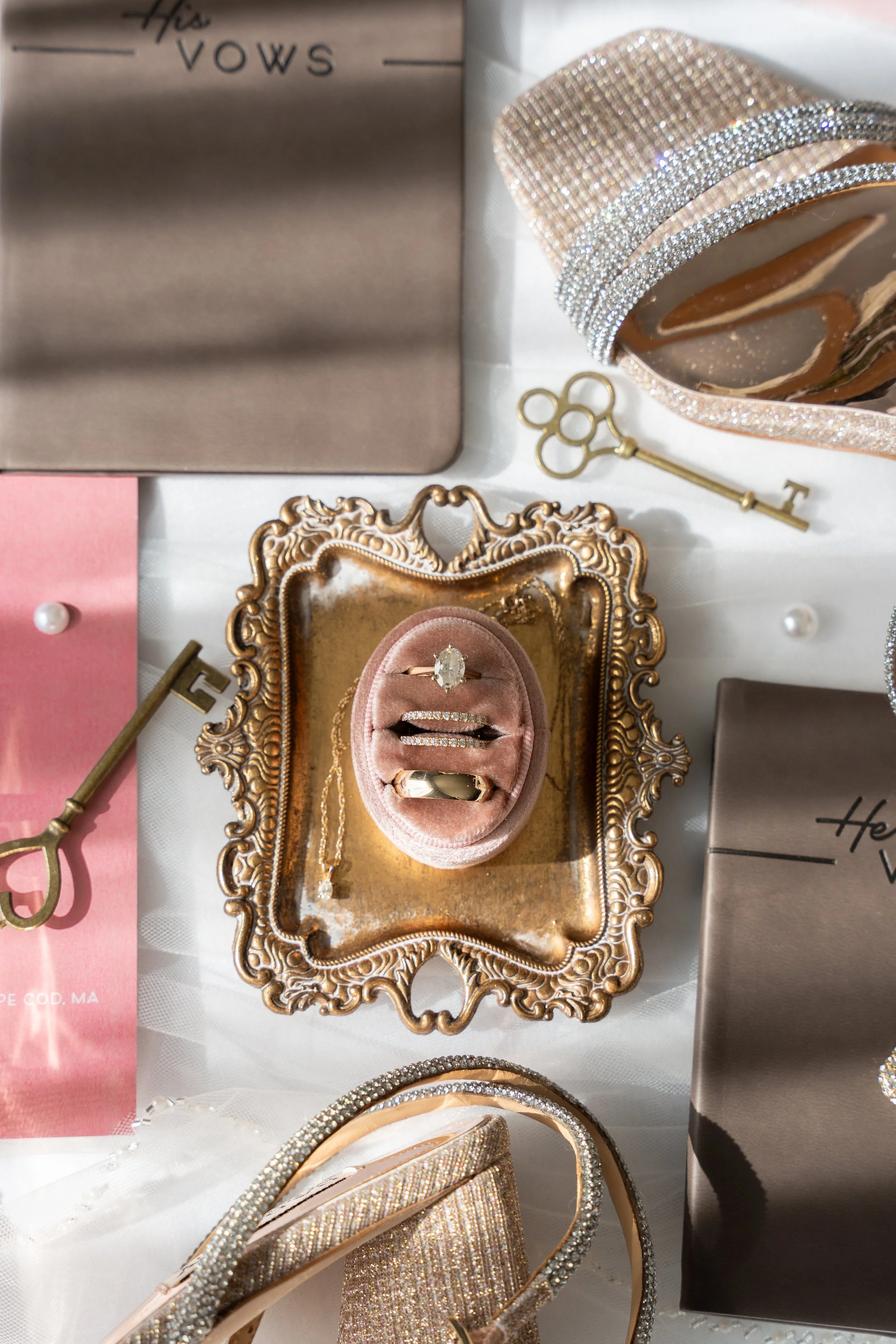 Display of jewelry and accessories on ornate tray, including rings, necklaces, and a bracelet, with a pink ring box, a gold key, and decorative boxes in the background during a wedding at Red Jacket Resort in Cape Cod. 