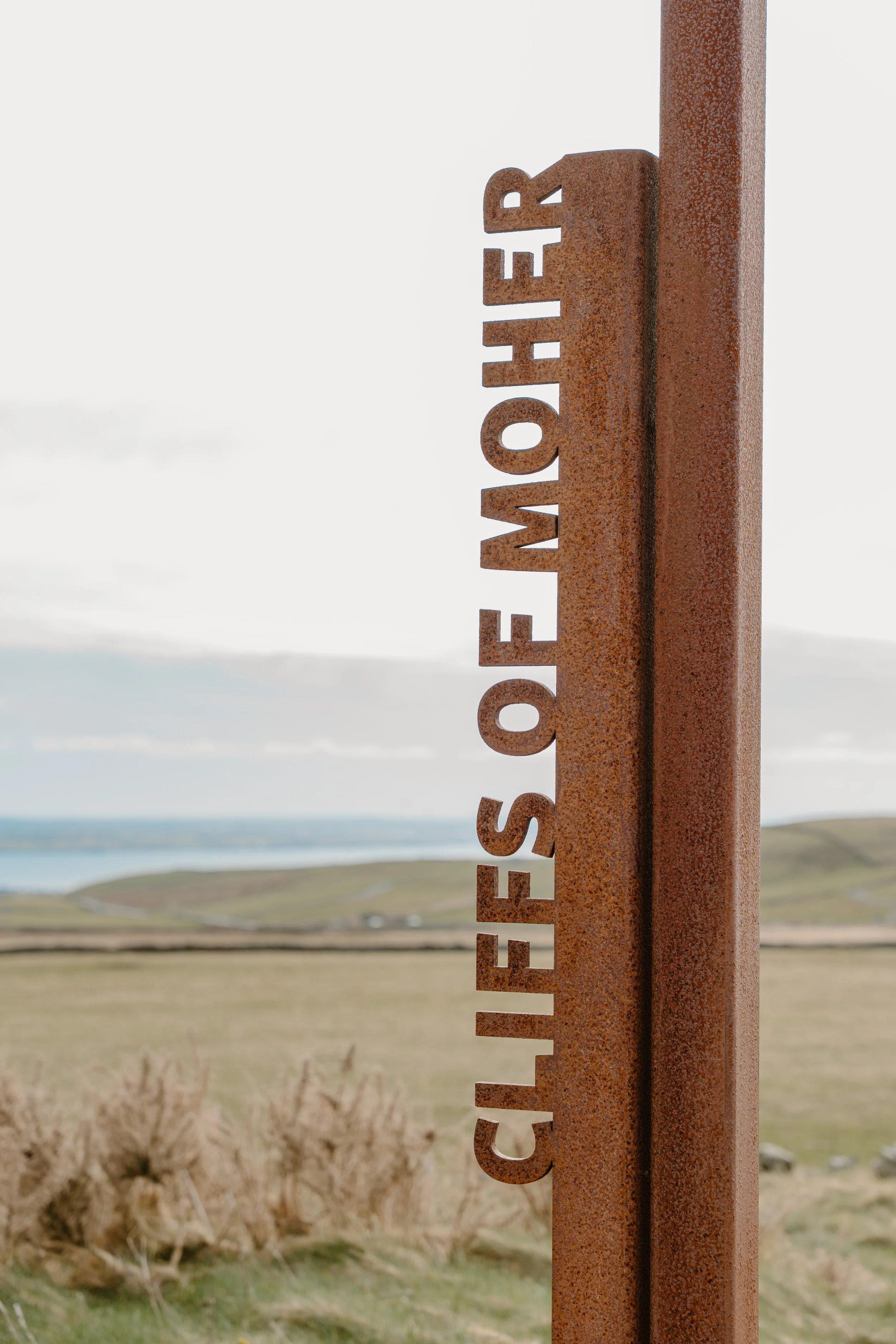 A rusted metal signpost with the words Cliffs of Moher cut out, standing in a grassy landscape with hills and a body of water in the background in Galway, Ireland. 