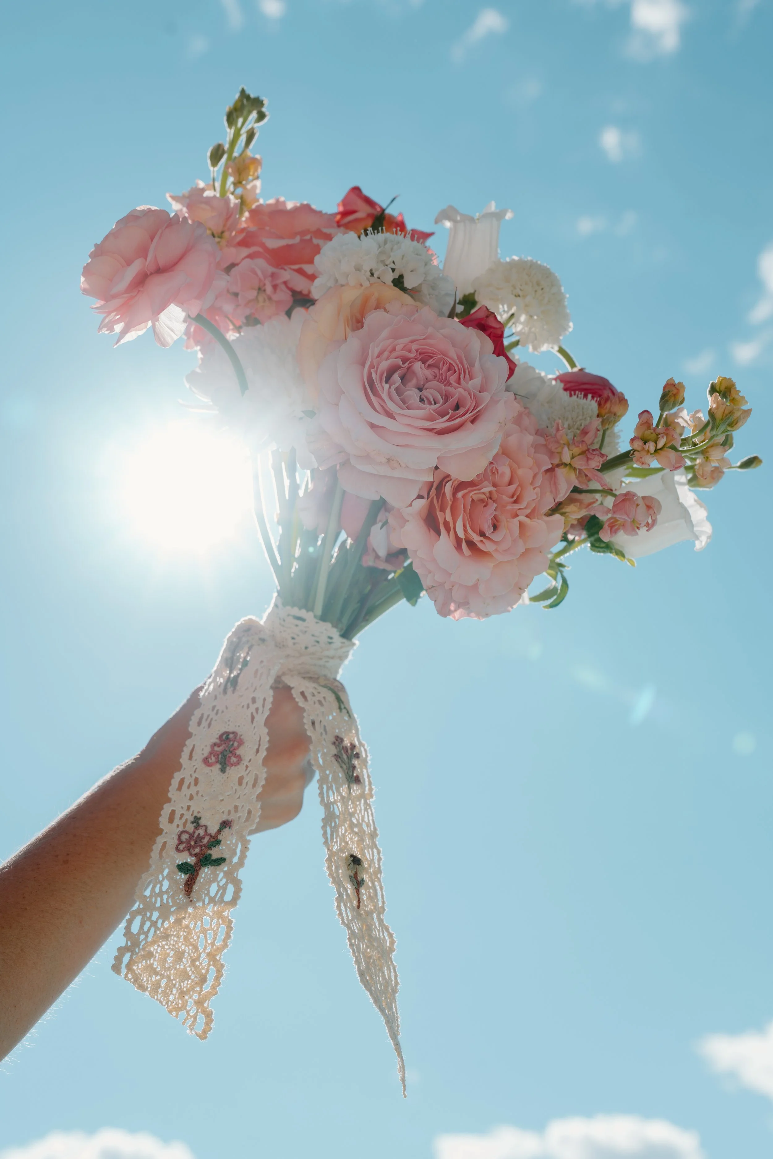 Person holding a bouquet of pink and white flowers against a clear blue sky with the sun shining behind the bouquet at a wedding in Bath, Maine. 