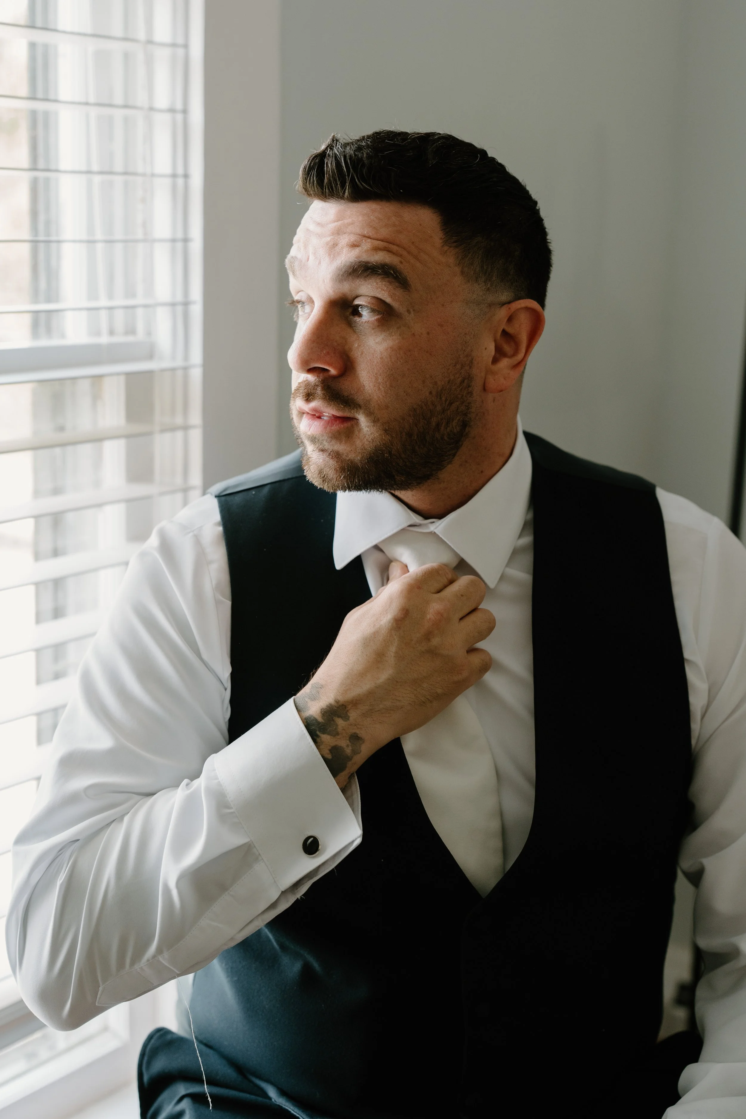 A man with short dark hair and facial hair, adjusting his tie while looking out a window with white blinds, dressed in a white shirt and black vest for a wedding at Holiday Hill Day Camp in Connecticut.