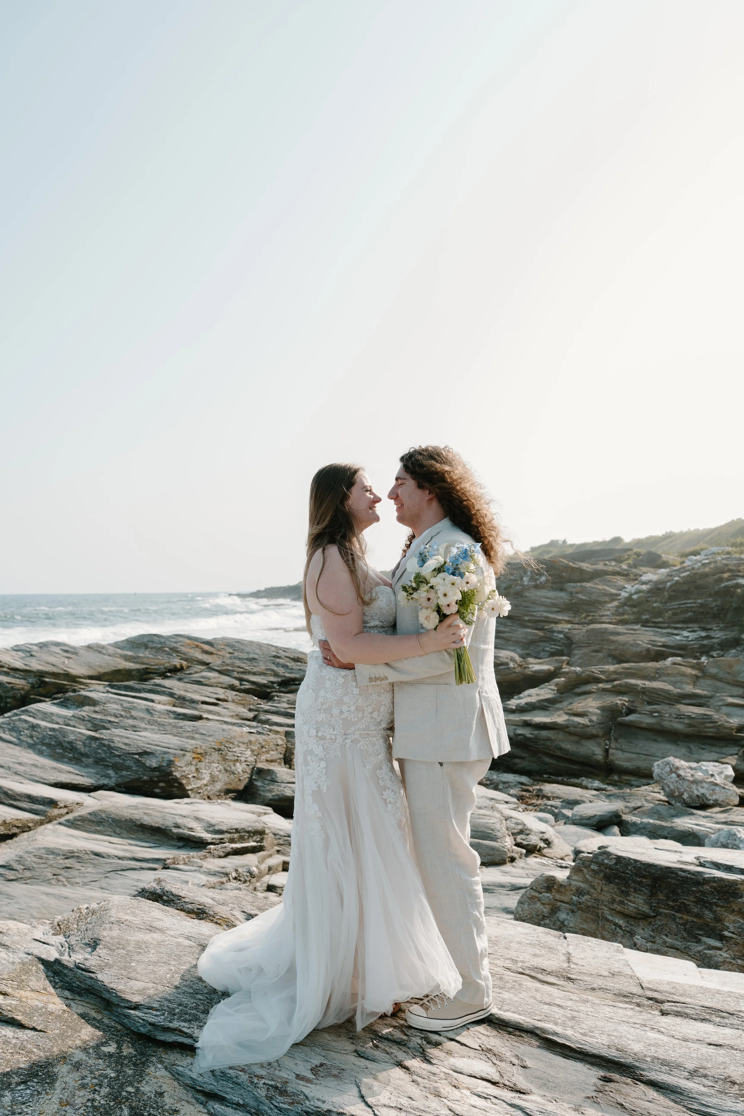 A couple in wedding attire sharing a moment on rocky beach with ocean and hills in background at an elopement at Beavertail State Park in Rhode Island. 