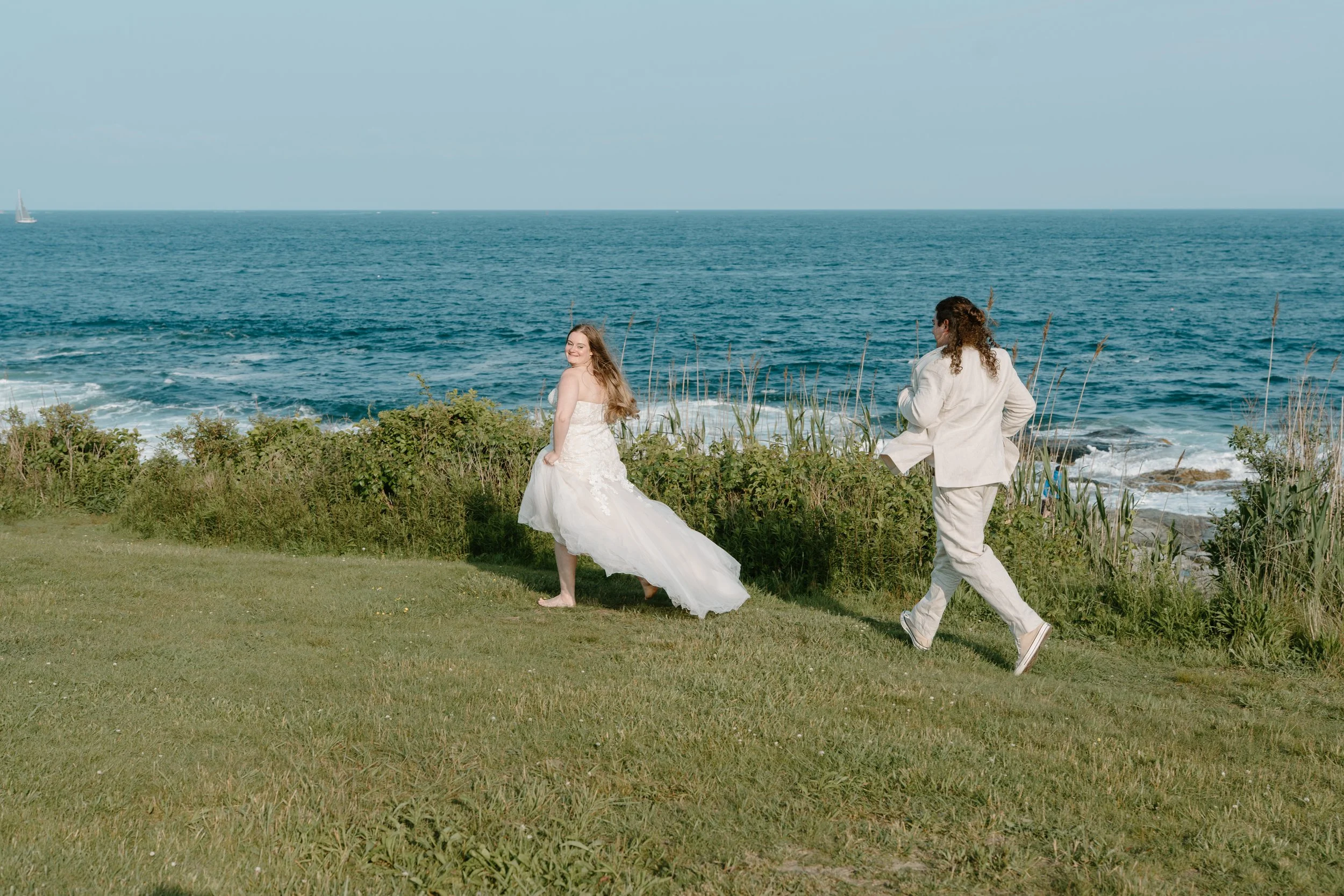 A bride in a white wedding dress smiling, and a groom in a white suit walking along a grassy area by the ocean with waves and sailboats in the distance at an elopement at Beavertail State Park in Rhode Island. 