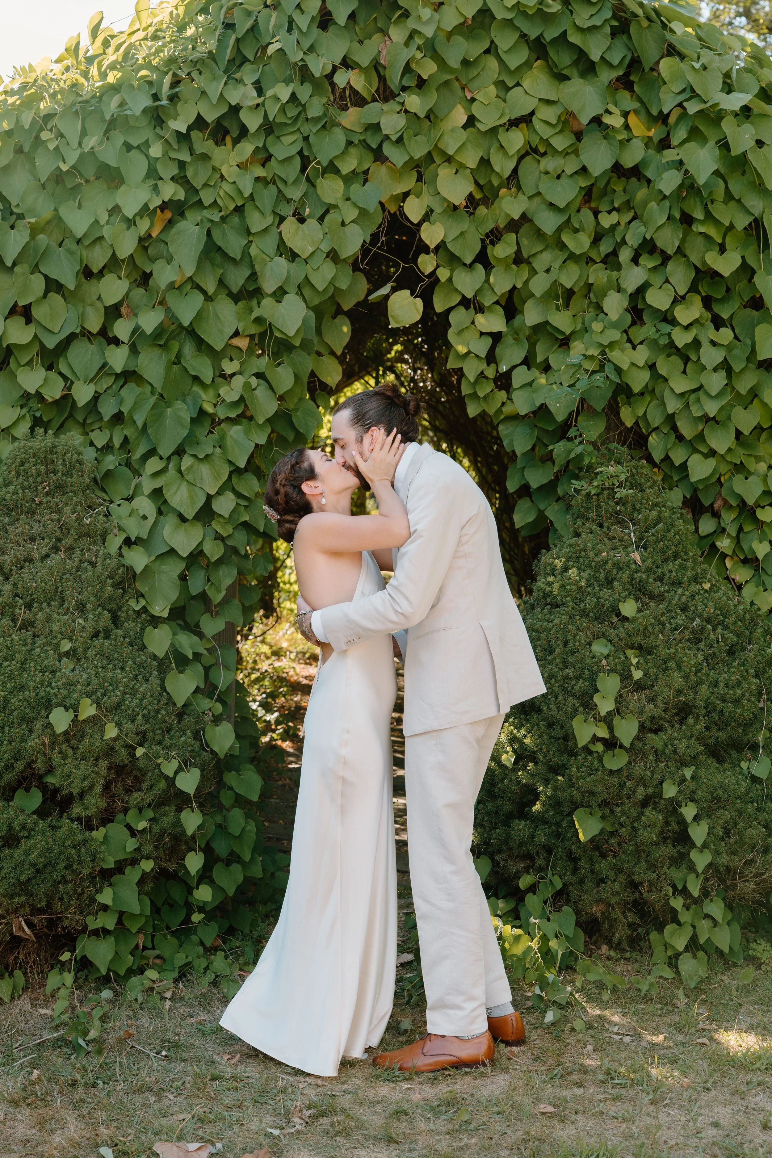 A couple dressed in white, sharing a kiss under a green leafy archway, surrounded by bushes on a sunny day at a wedding in Bath, Maine. 