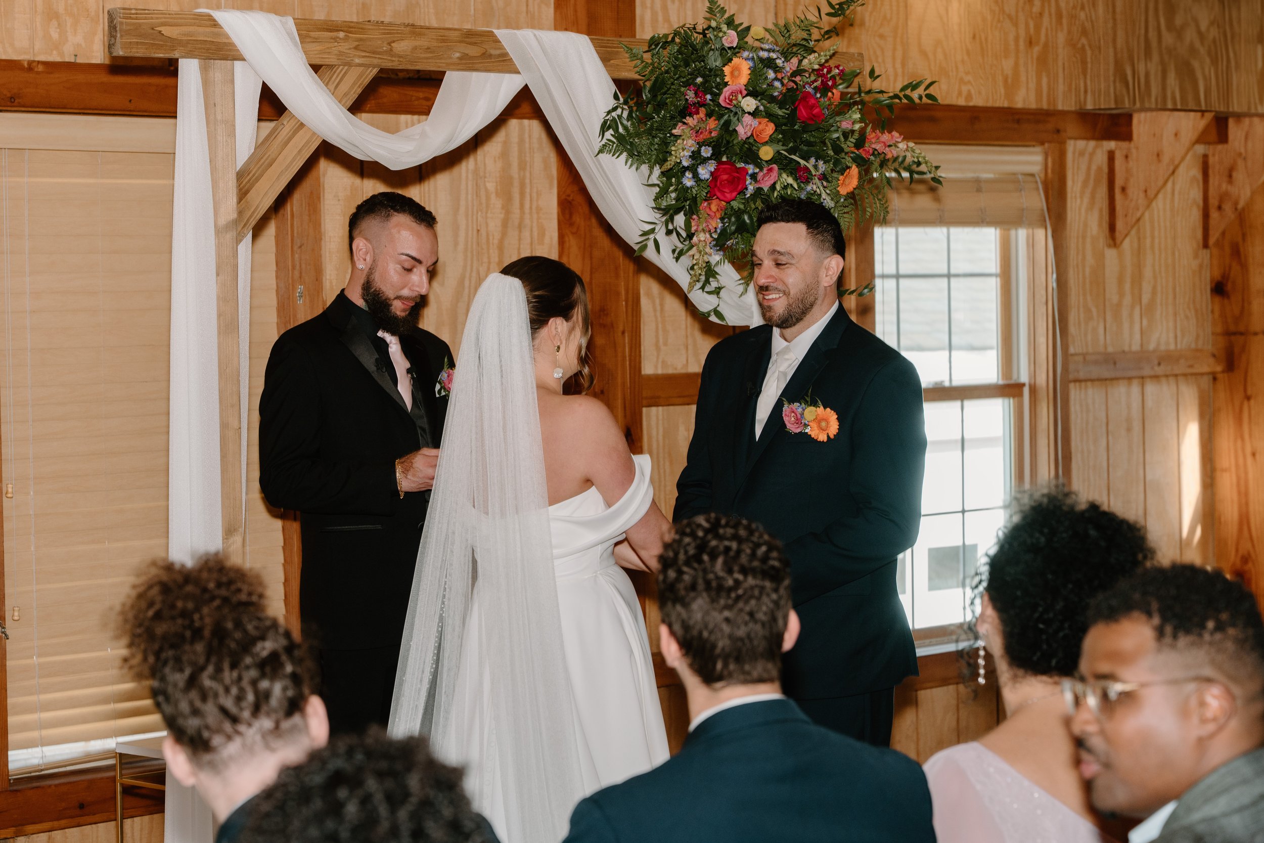 A wedding ceremony with a bride and groom exchanging vows in a wooden venue, decorated with a floral arch and white drapery, while guests observe for a wedding at Holiday Hill Day Camp in Connecticut.
