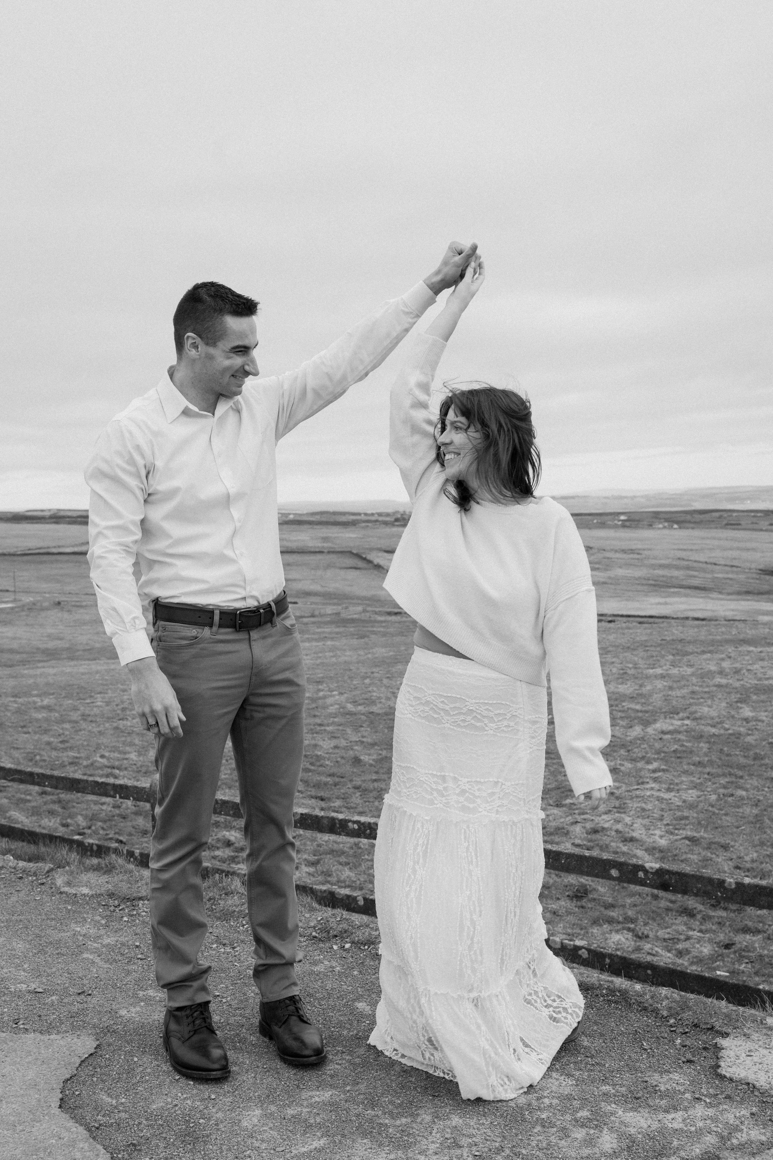 A man and woman dancing outdoors on a cloudy day, holding hands, smiling, with open landscape in the background for an engagement photo session in Galway, Ireland. 