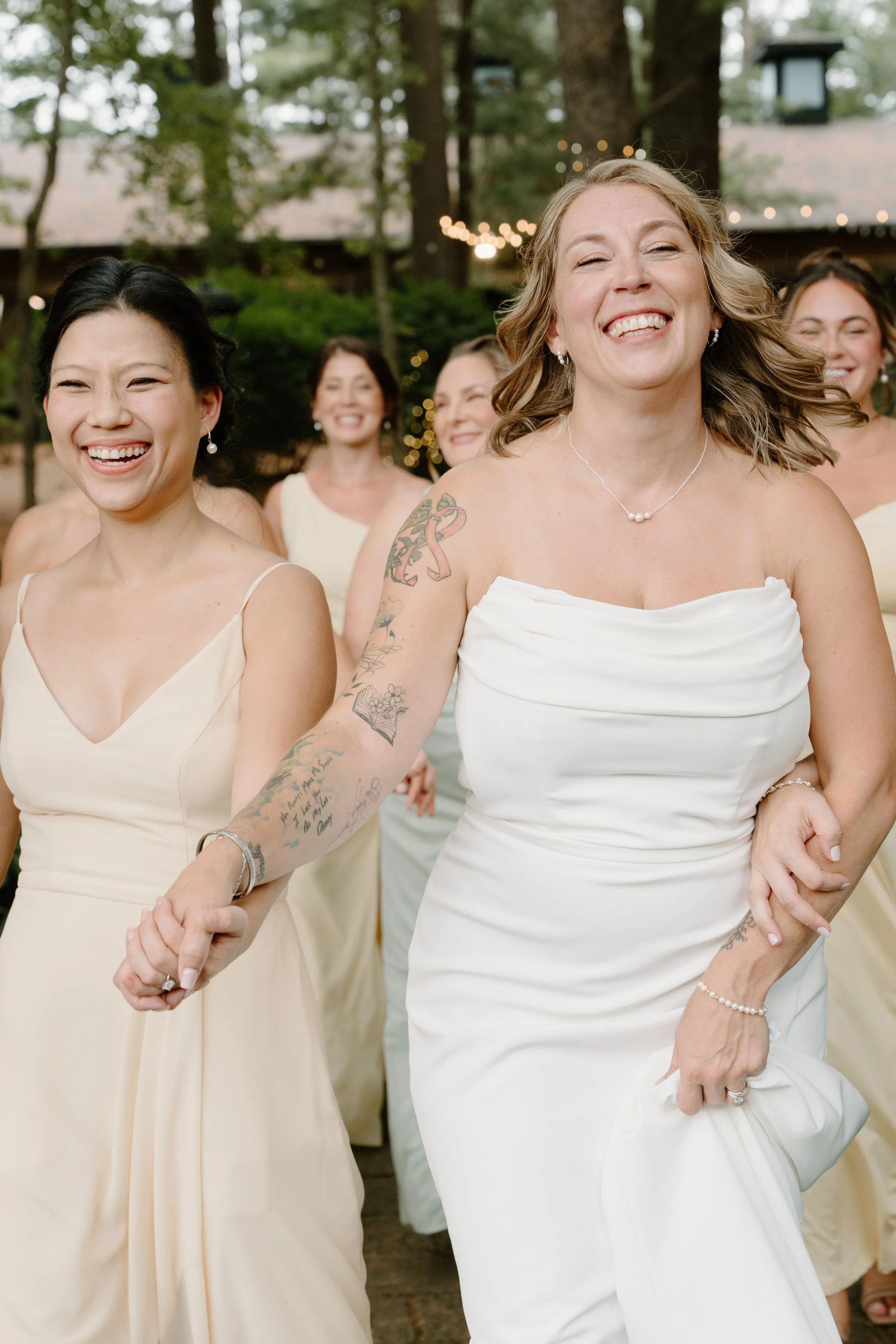 A group of women, some with tattoos, smiling and holding hands at an outdoor celebration, with string lights and trees in the background. One woman in the foreground wears a strapless white dress for a wedding at Tunxis Country Club in Connecticut.