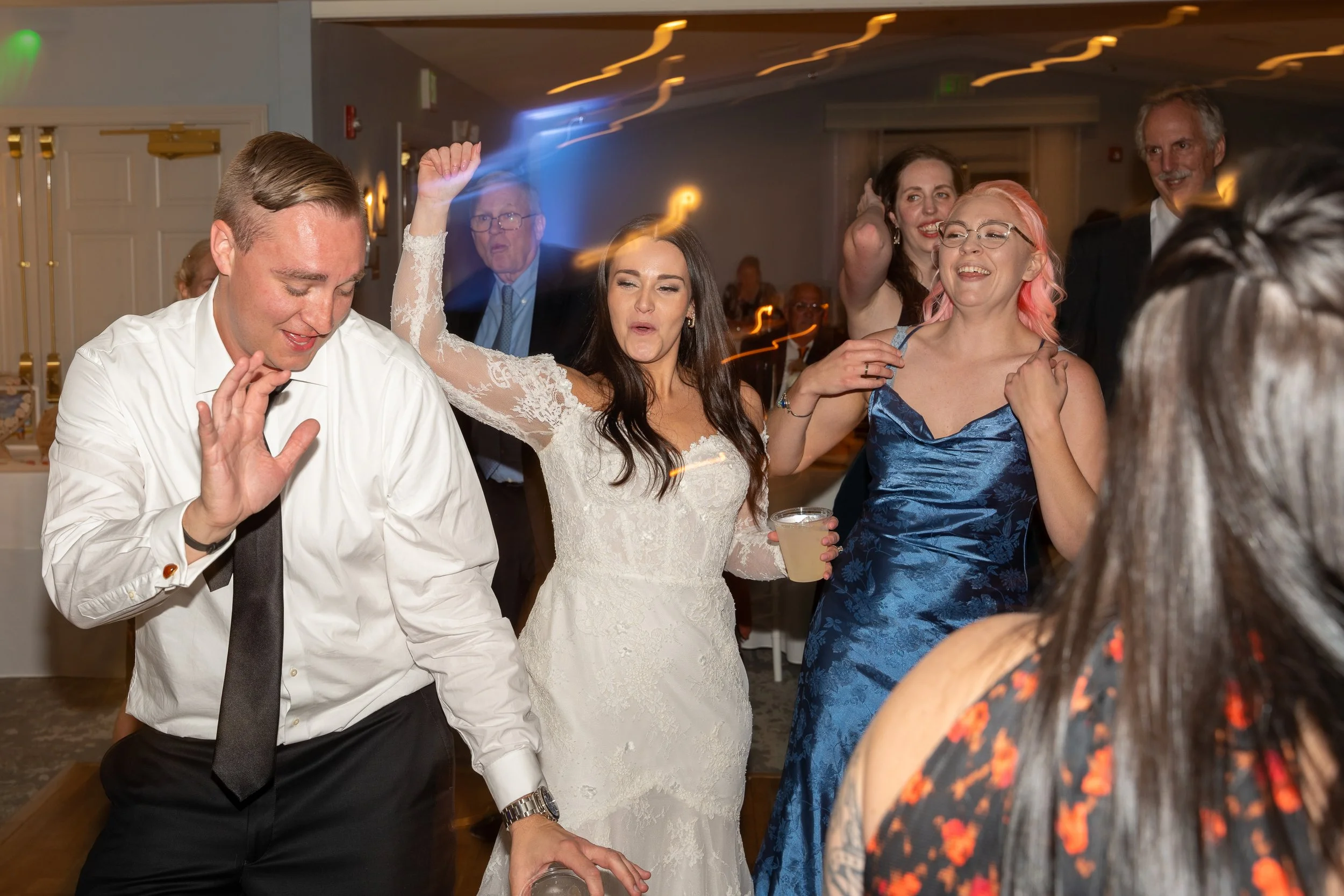 People dancing and enjoying themselves at a celebration or wedding reception, with a woman in a white lace dress, a man in a white shirt and black tie, and several women in colorful dresses during a wedding at Red Jacket Resort in Cape Cod. 