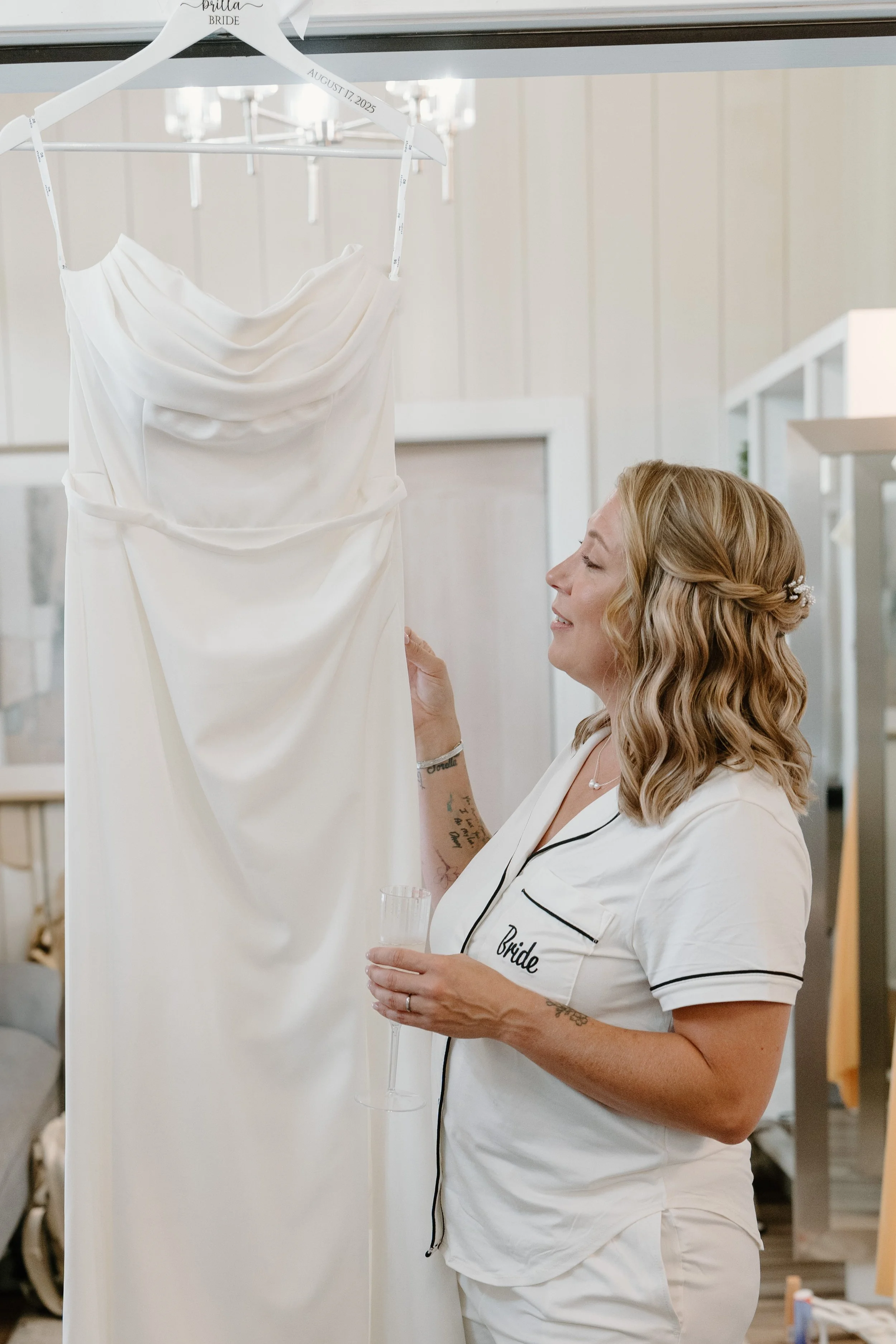 A woman with wavy blonde hair is standing next to a white dress hanging on a hanger. She is holding a glass of champagne and smiling at the dress for a wedding at Tunxis Country Club in Connecticut. 