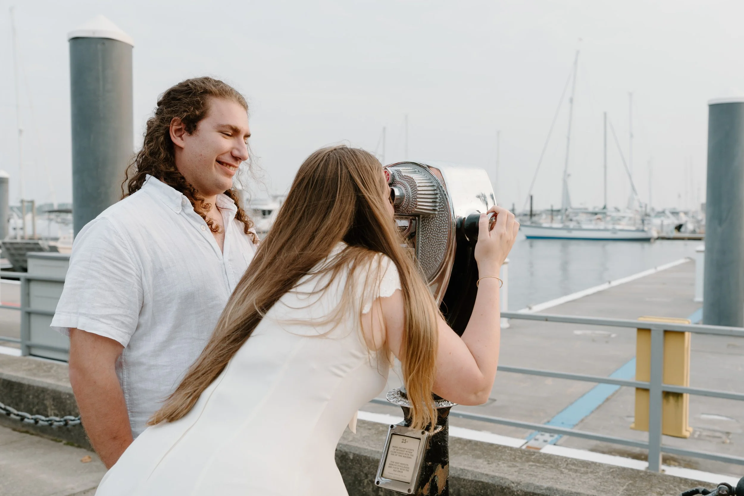 A couple by the marina looking through a coin-operated viewing telescope, with boats in the background after an elopement in Newport, Rhode Island. 