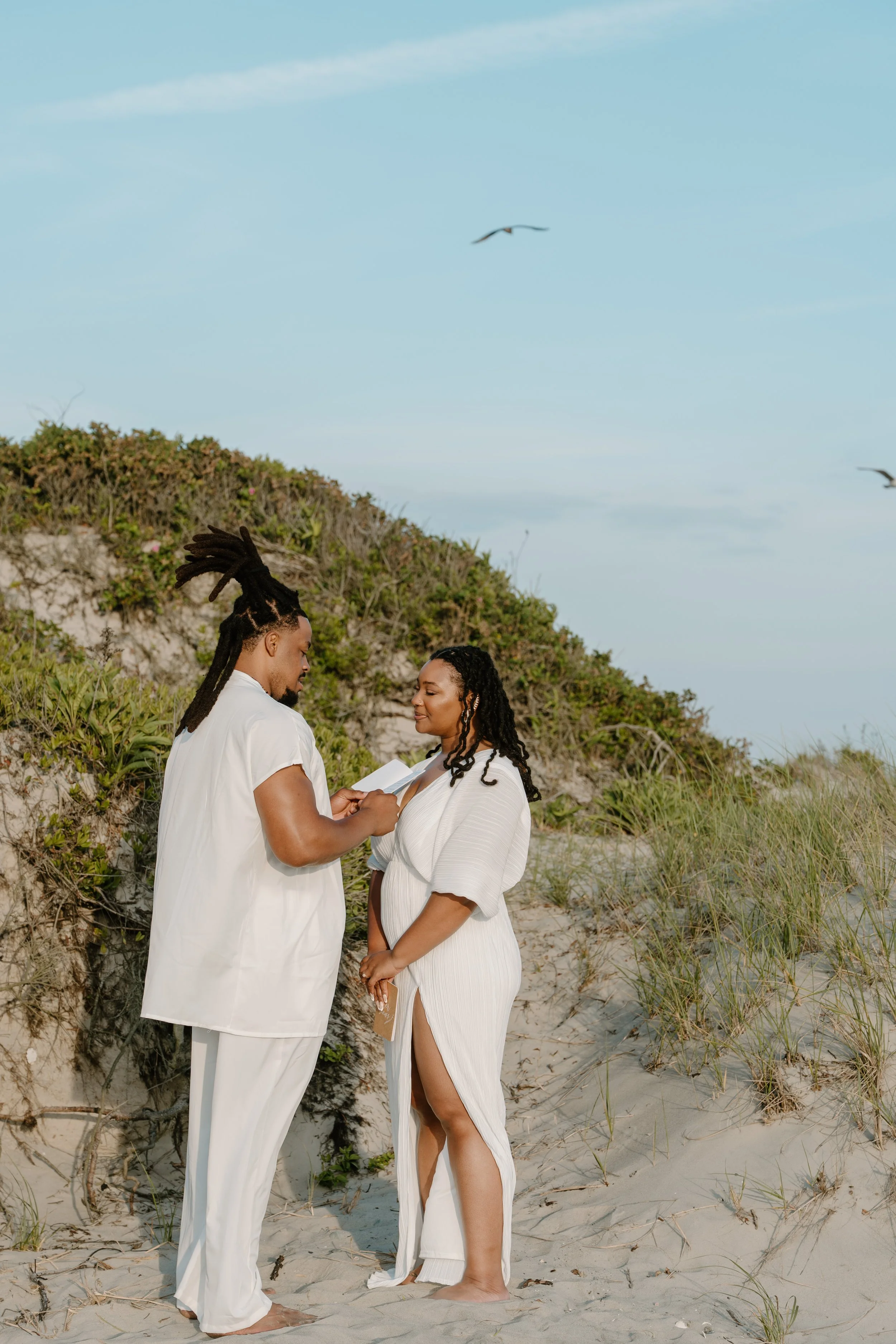 A couple wearing white, standing on beach sand, with greenery and sand dunes behind them, exchanging vows with seagulls flying overhead at an elopement at Second Beach in Rhode Island. 