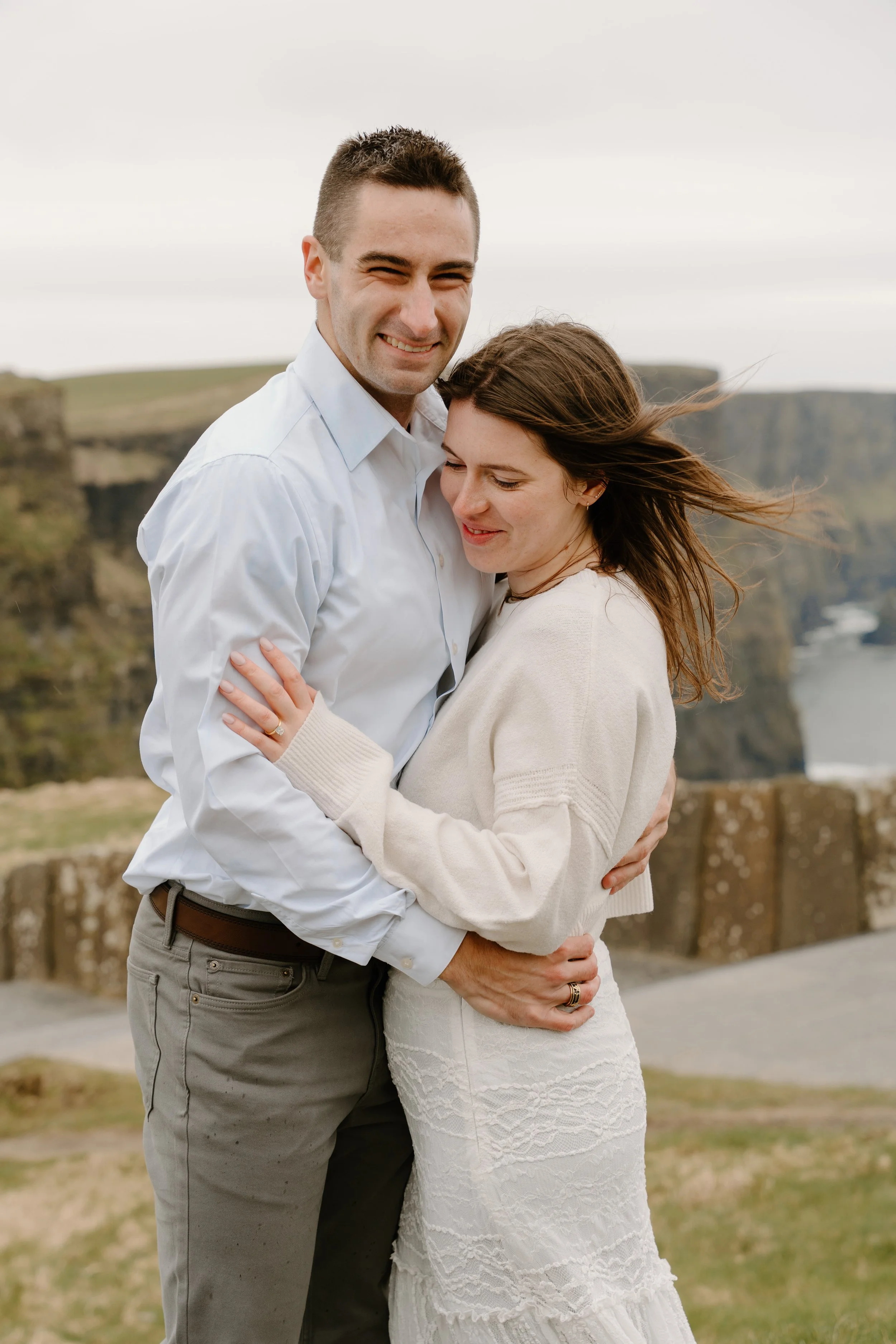 A smiling couple hugging outdoors with cliffs and a body of water in the background for an engagement photo session in Galway, Ireland. 