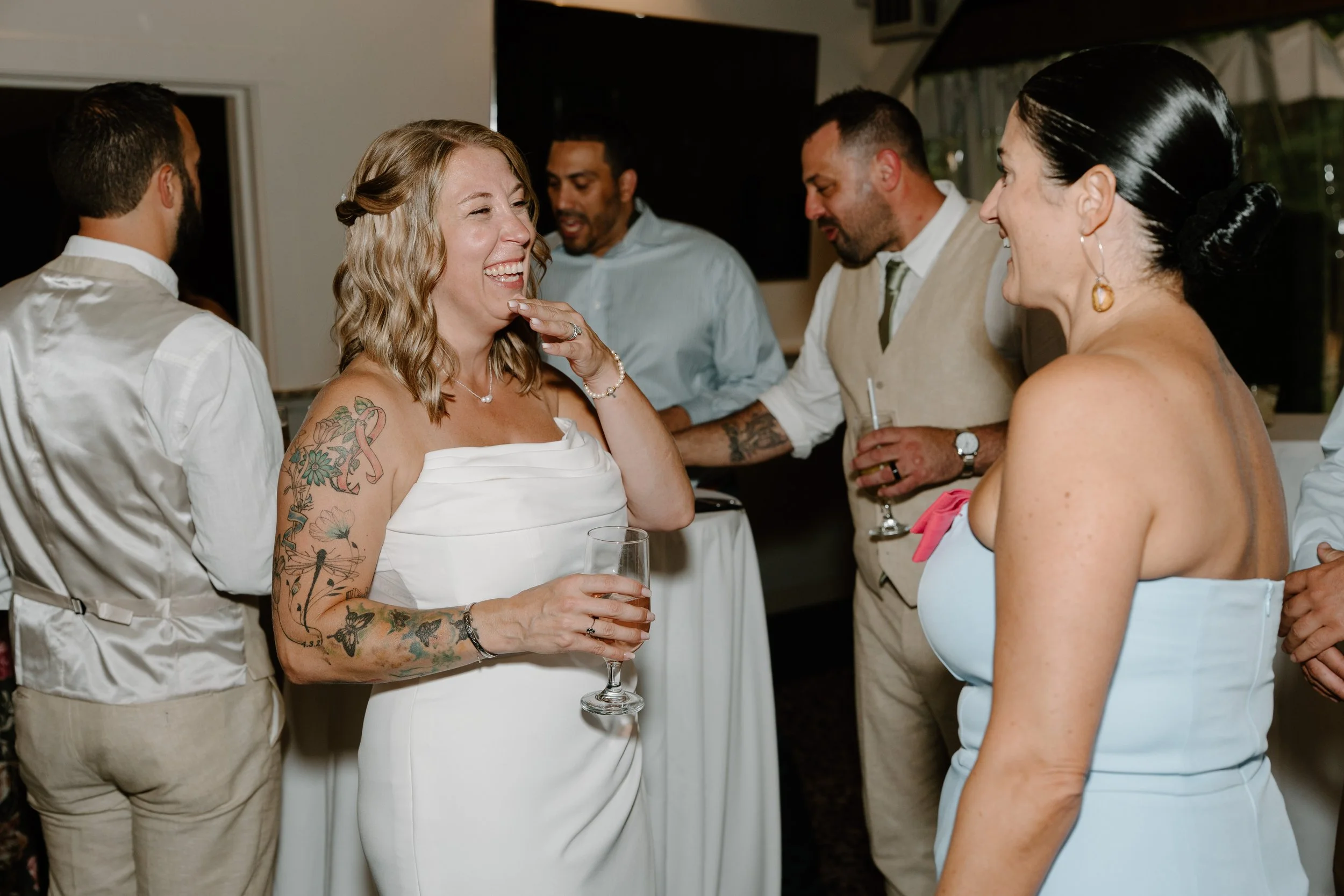 A group of people at a celebration or party, with one woman in a white strapless dress laughing and holding a glass of wine, surrounded by others who are talking and smiling for a wedding at Tunxis Country Club in Connecticut. 