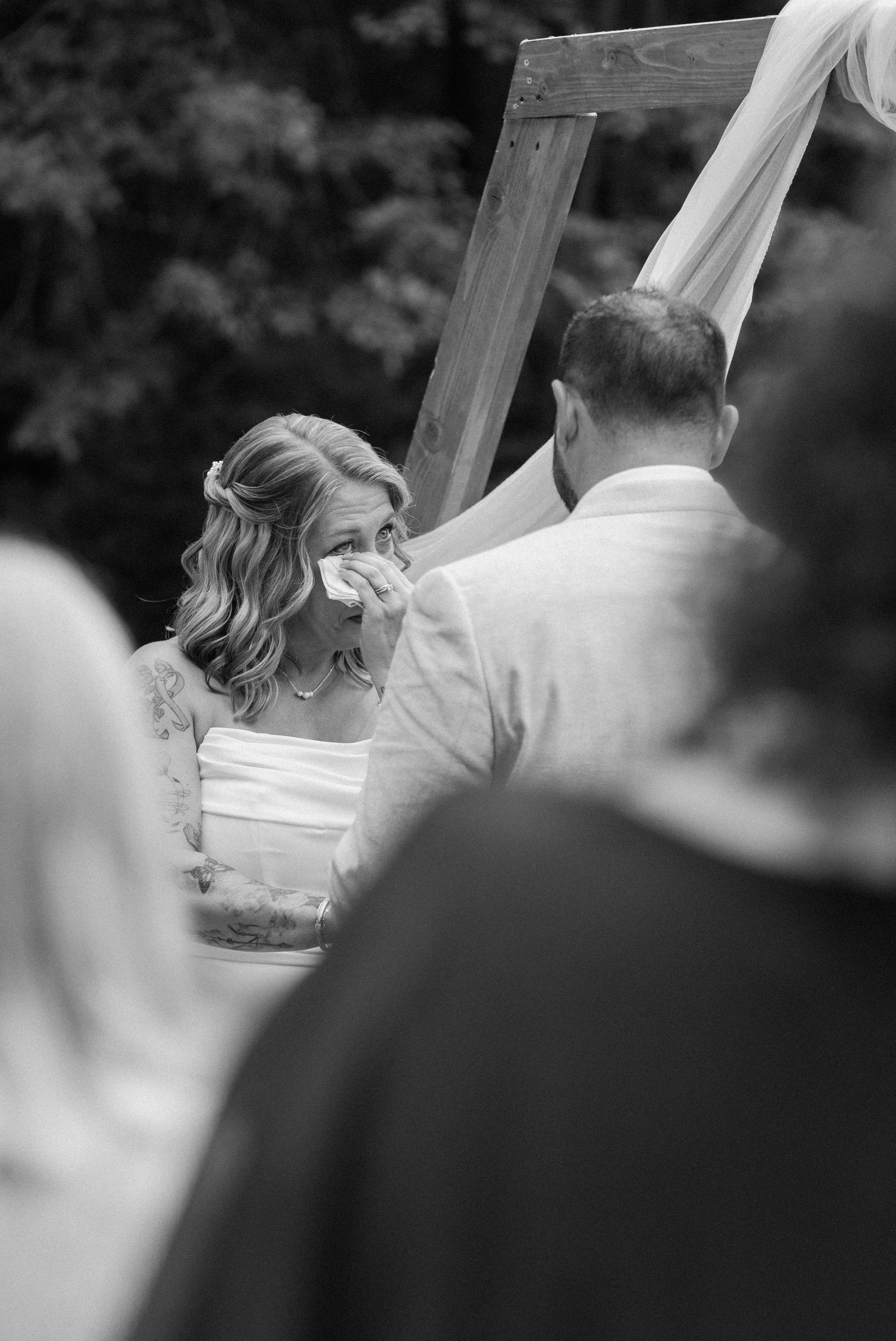 A woman in a wedding dress is crying as she holds a tissue, during her wedding ceremony, with a man facing her. The scene is outdoors with blurred greenery in the background for a wedding at Tunxis Country Club in Connecticut. 