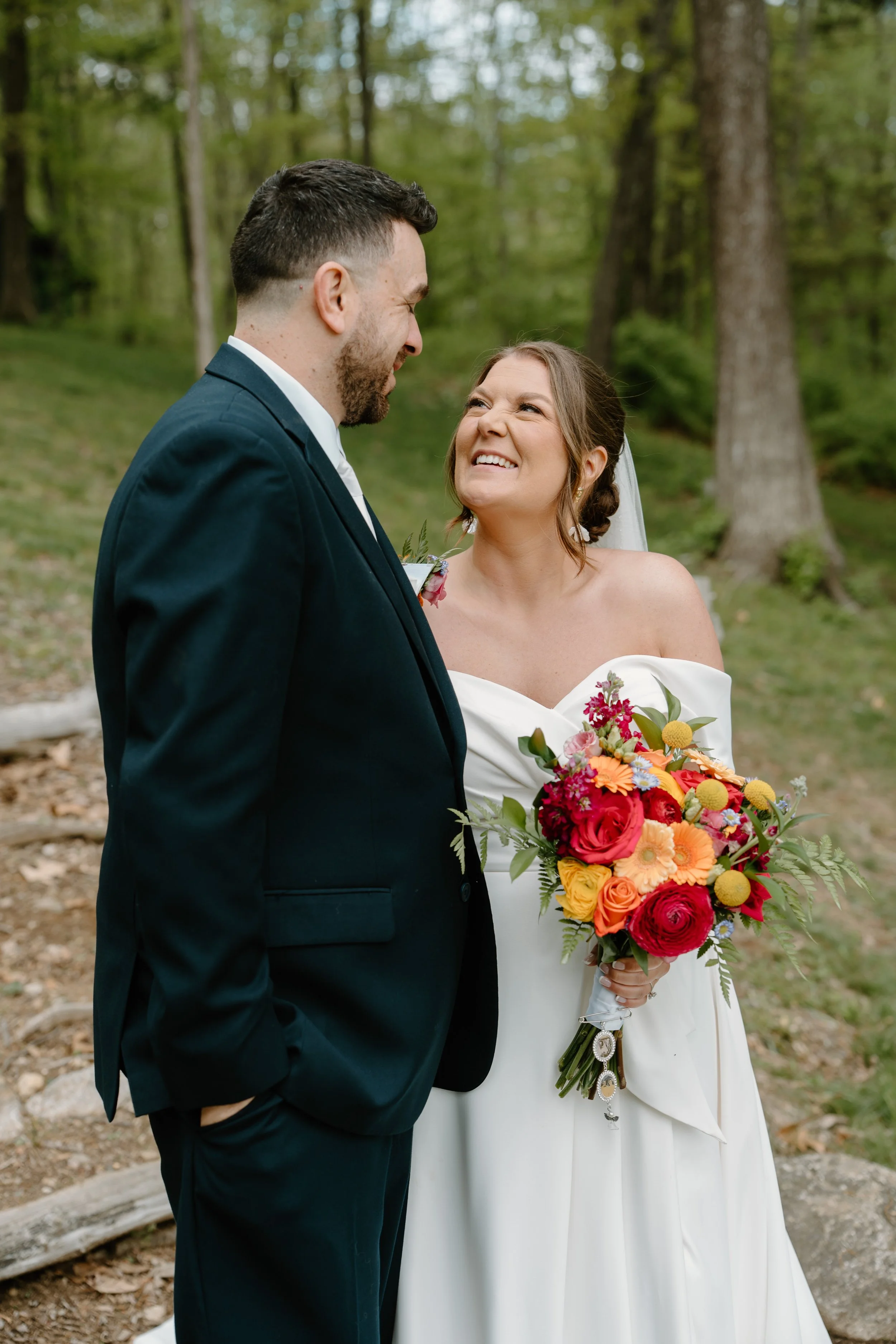 A bride and groom share a joyful moment in a forested outdoor setting, with the bride holding a colorful bouquet and the groom in a dark suit for a wedding at Holiday Hill Day Camp in Connecticut.