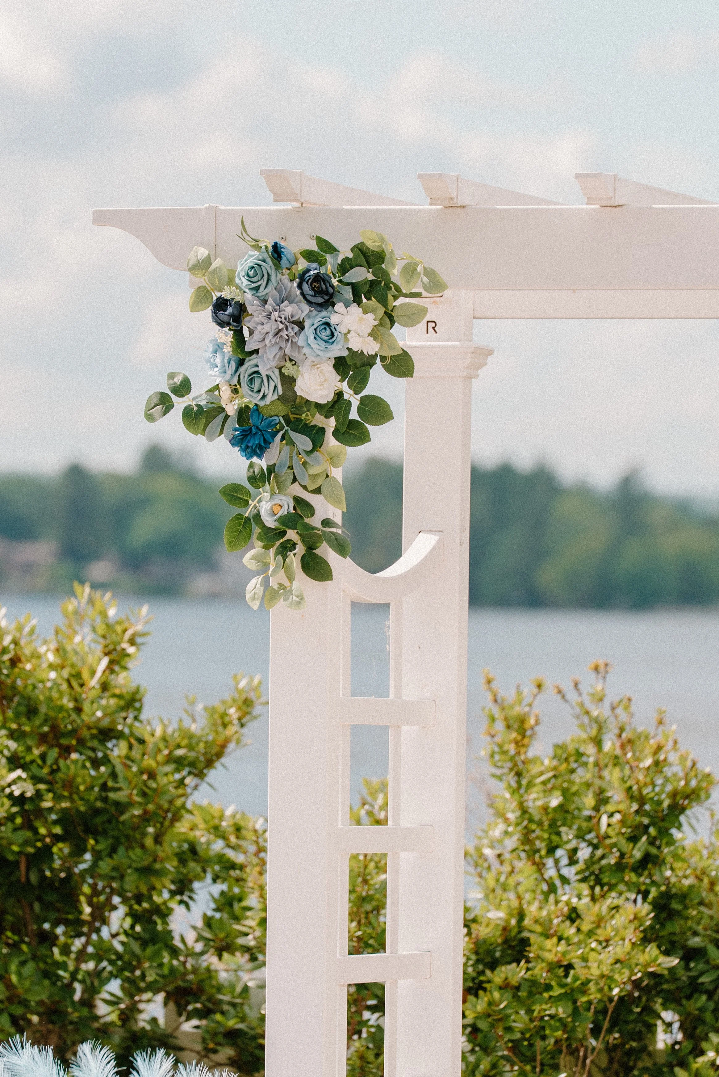White pergola with blue and white flower arrangement against a lake and greenery backdrop.