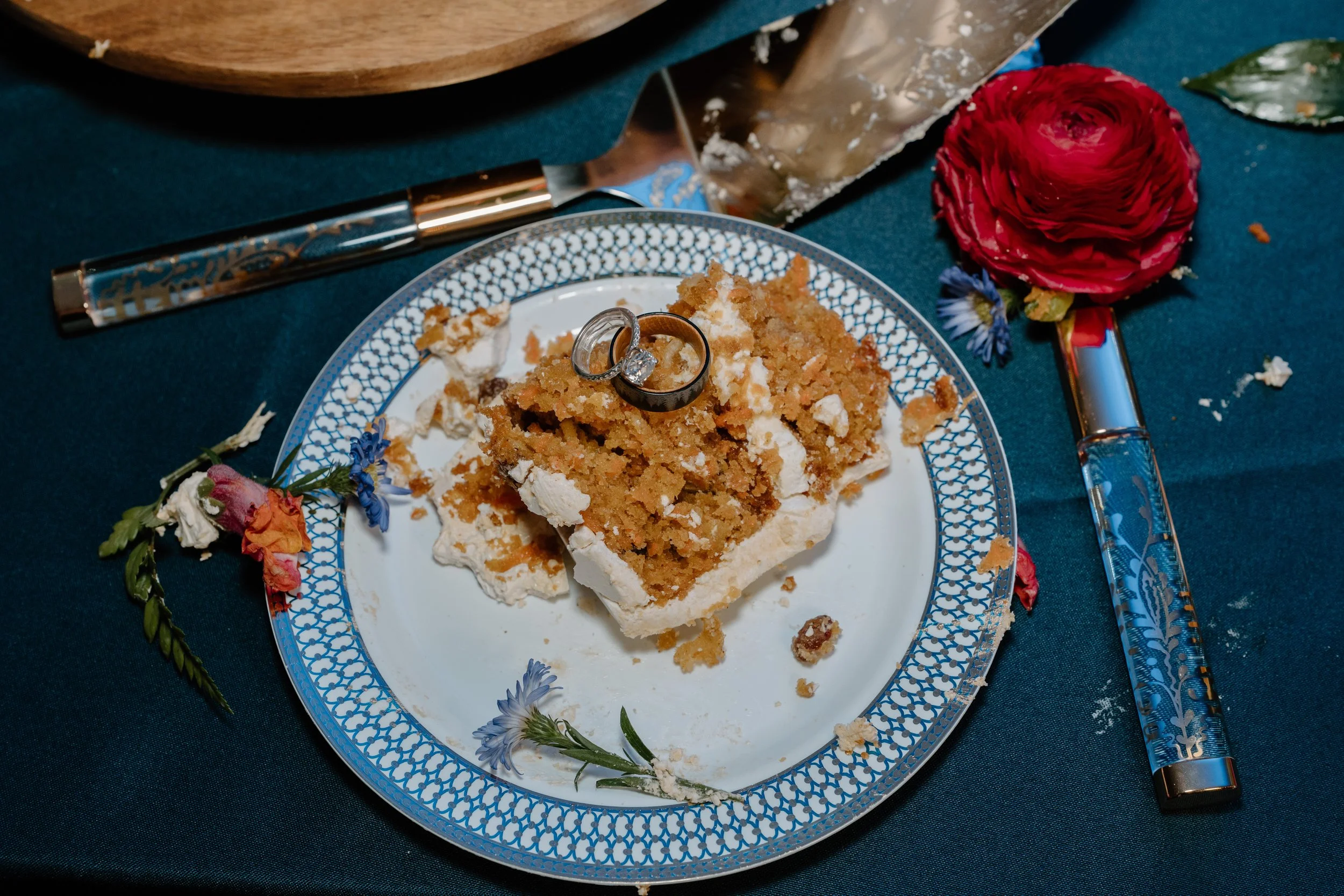 A partially eaten piece of cake with white icing and crumb topping on a decorative plate, with a wedding ring and engagement ring on top, surrounded by flowers, on a teal tablecloth for a wedding at Holiday Hill Day Camp in Connecticut.