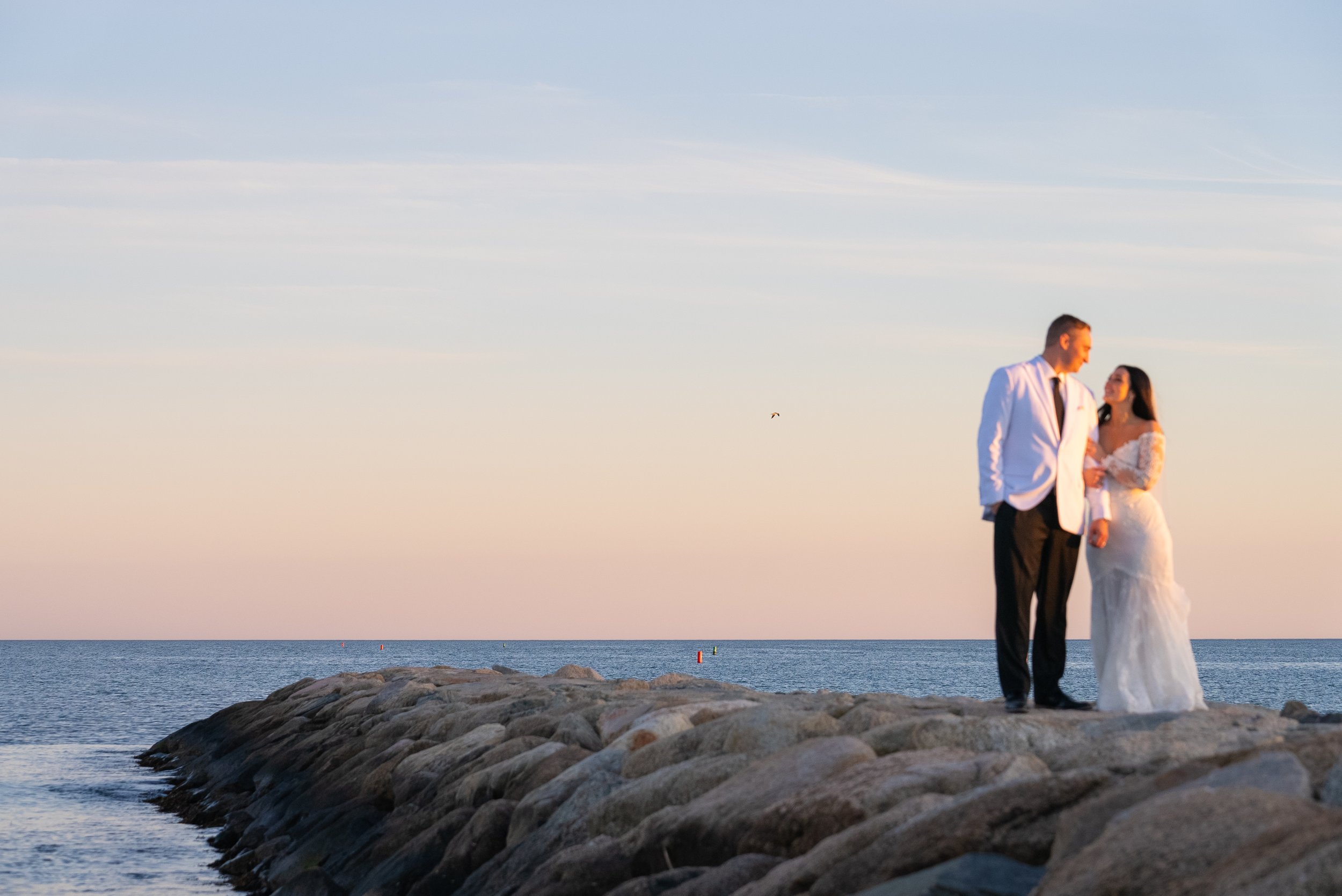 A couple dressed in formal wedding attire walking on a rocky pier by the water at sunset, with a clear sky and calm ocean in the background during a wedding at Red Jacket Resort in Cape Cod. 