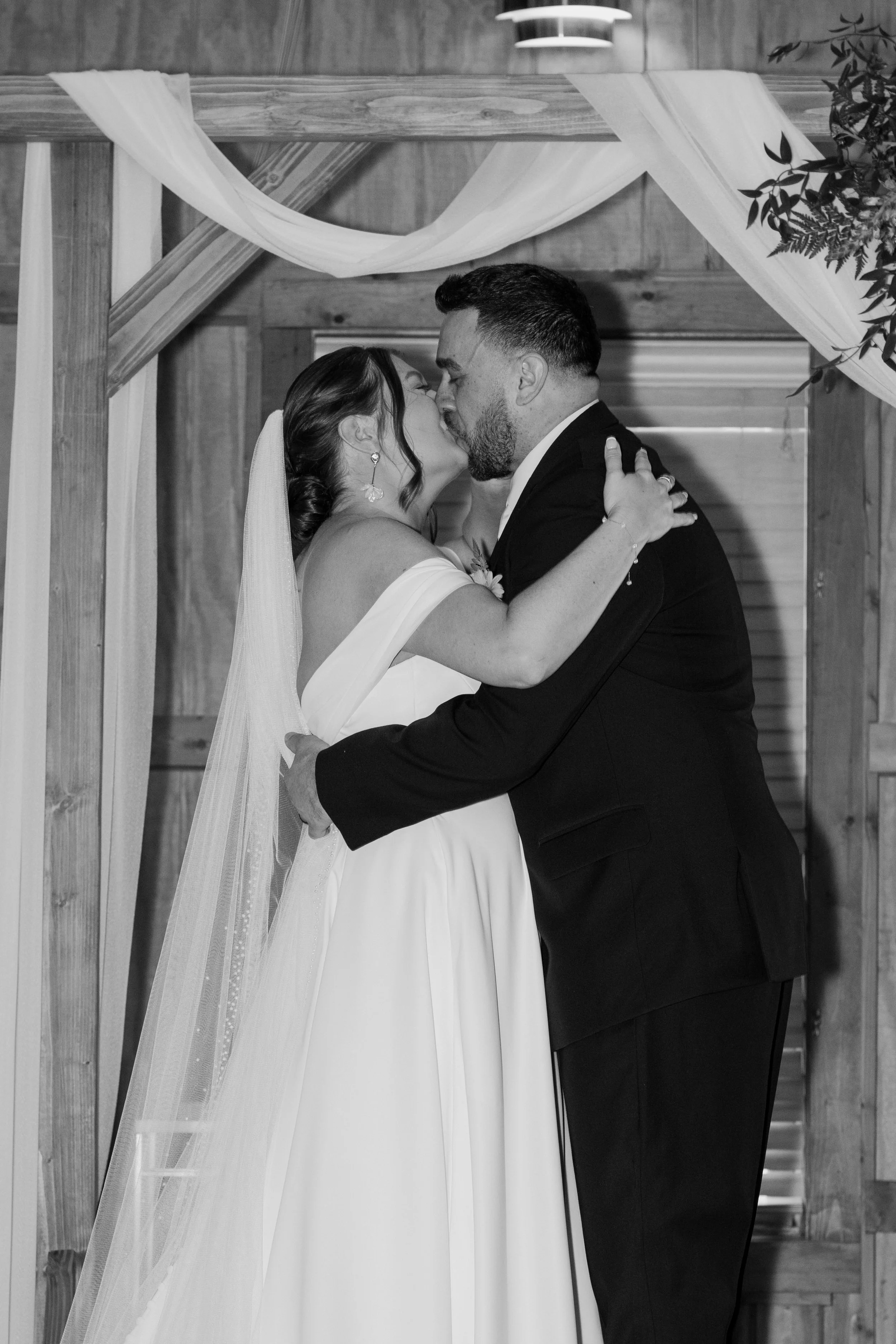 A bride and groom kissing during their wedding ceremony, standing under a wooden arch decorated with drapery and greenery for a wedding at Holiday Hill Day Camp in Connecticut.