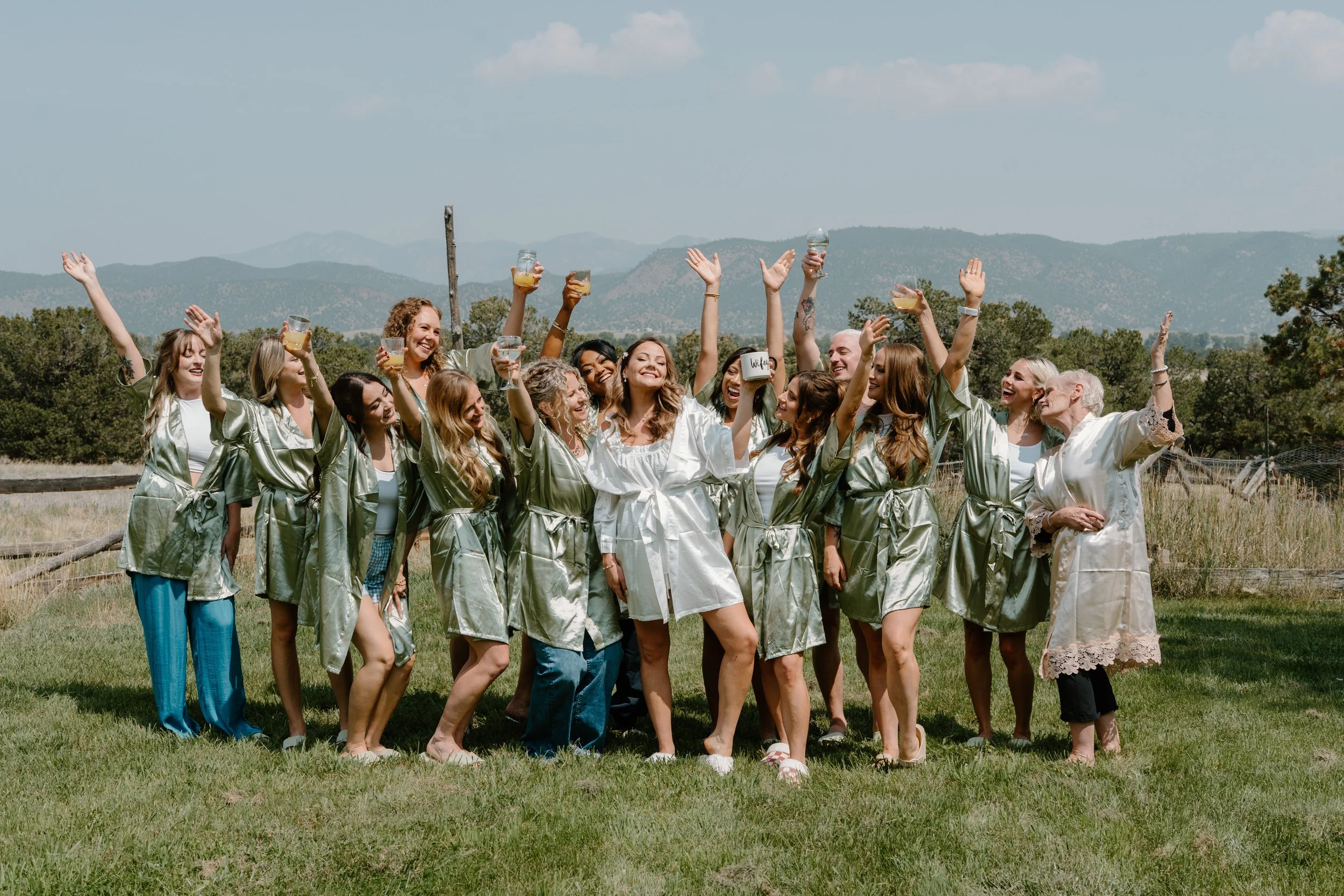 A group of women celebrating outdoors on a sunny day, wearing matching satin robes, with some holding drinks, standing on grass with mountains in the background at a wedding at Everett Ranch in Salida, Colorado.