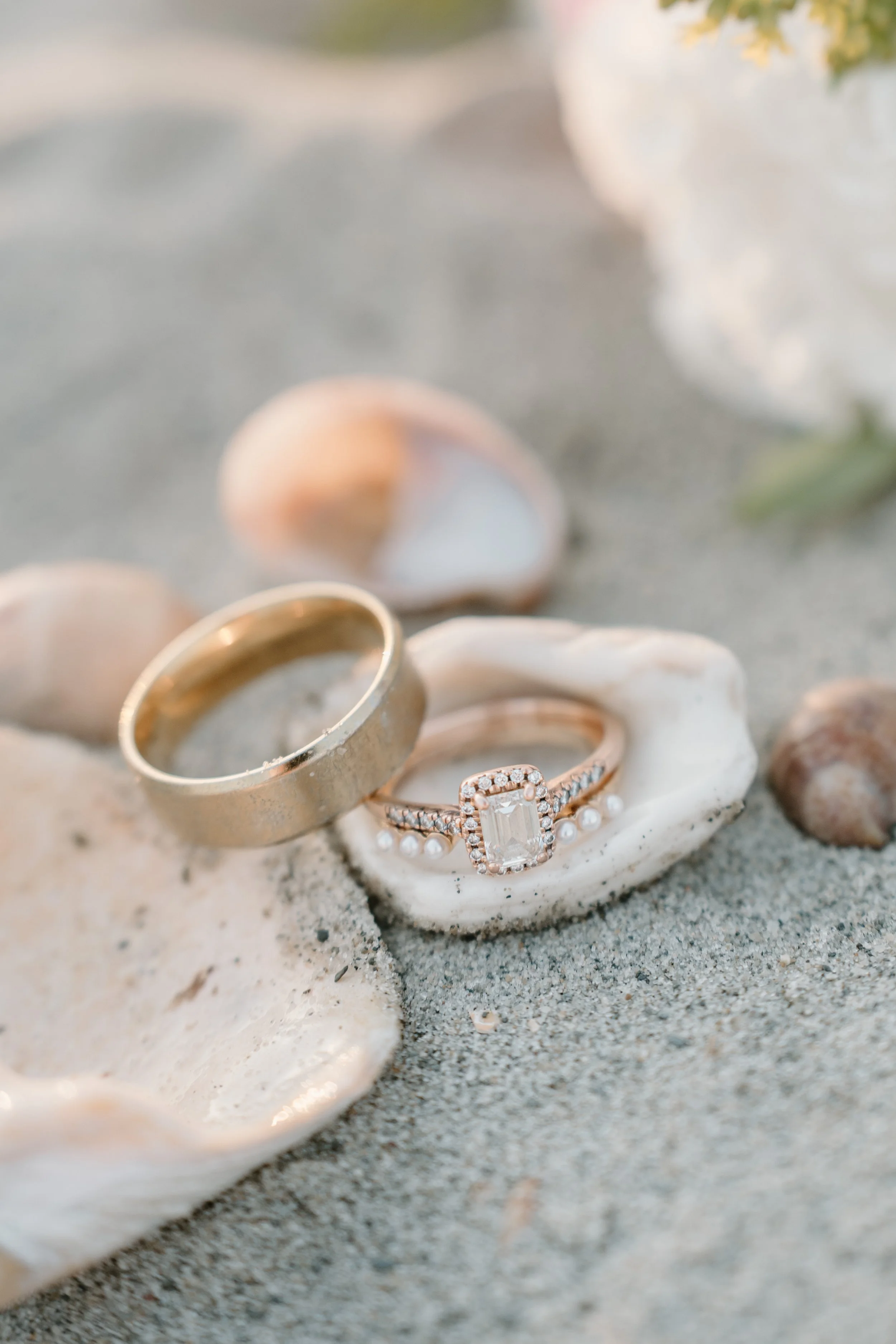 Two rings, one gold band and one with a stone and diamonds, resting on a seashell on the sand, with other seashells in the background at an elopement at Second Beach in Rhode Island. 