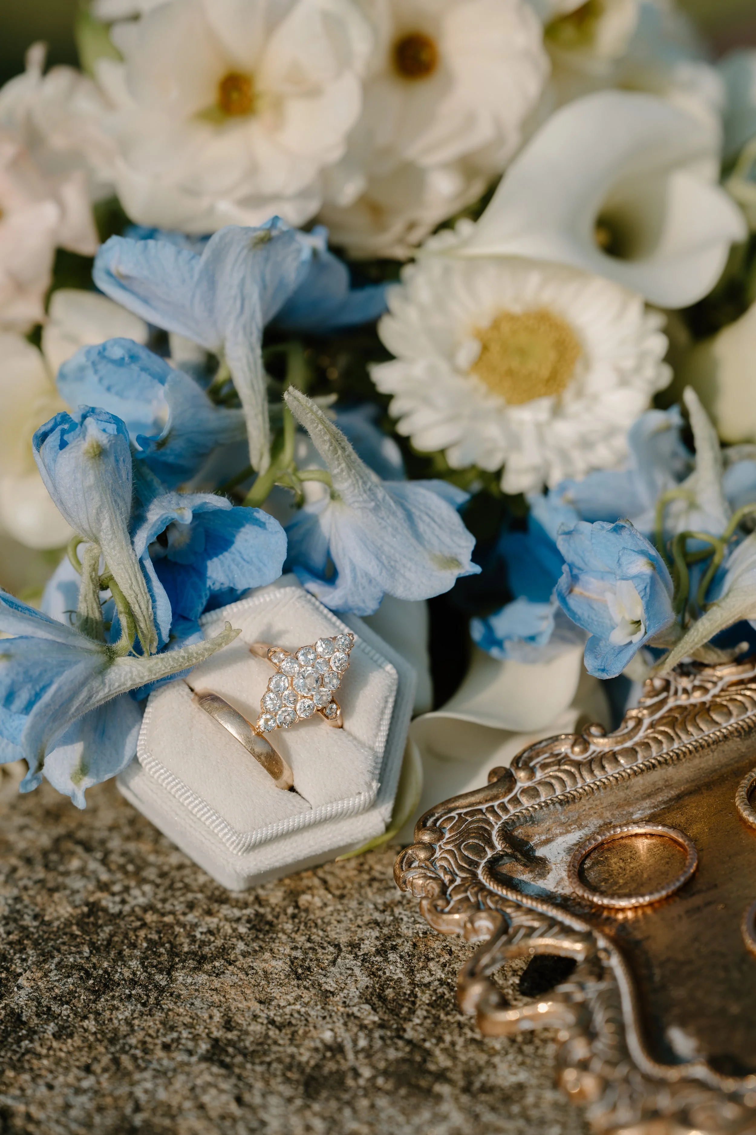 A diamond ring in a white velvet ring box, surrounded by blue and white flowers, next to an ornate gold tray at an elopement at Beavertail State Park in Rhode Island. 
