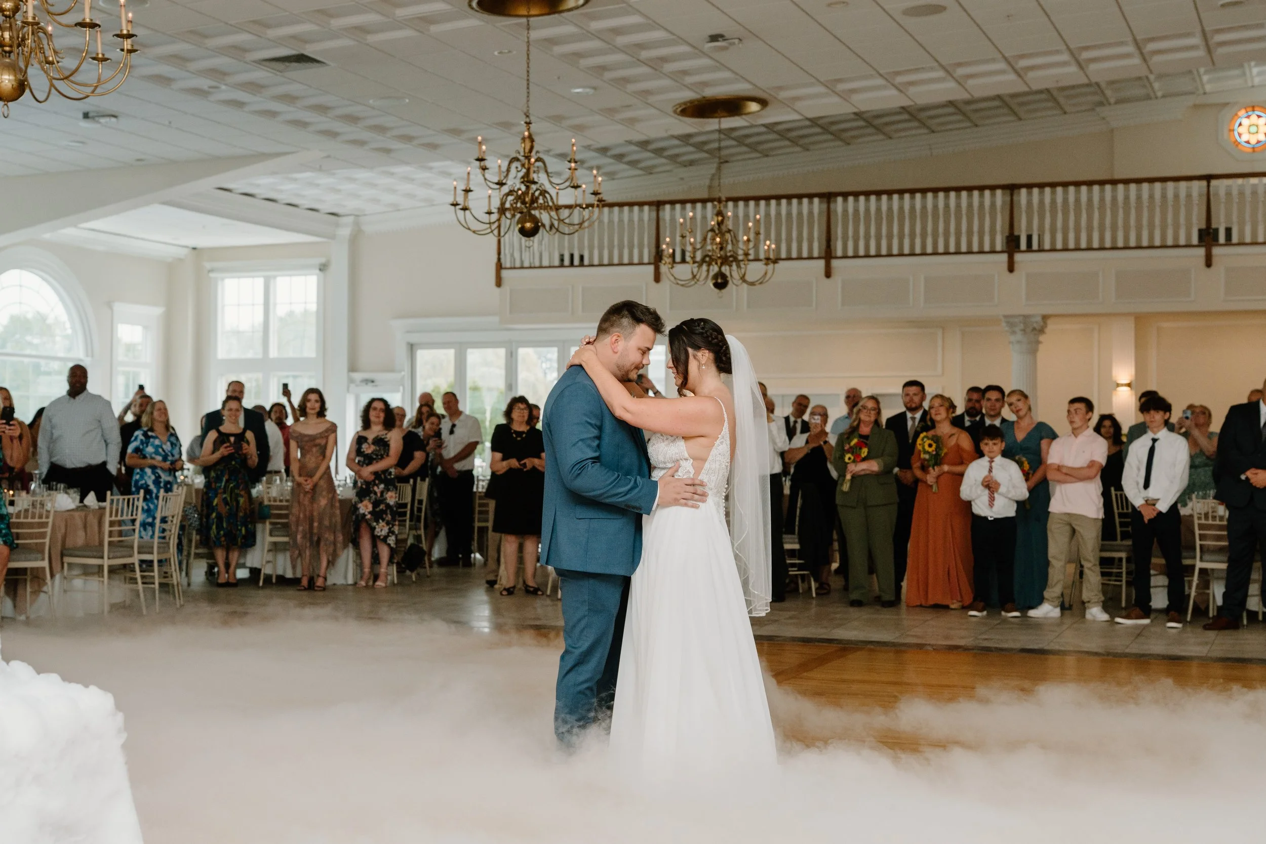 A bride and groom sharing their first dance at their wedding reception, surrounded by guests in a decorated banquet hall with chandeliers and large windows during a wedding at the Aqua Turf Club in Connecticut. 