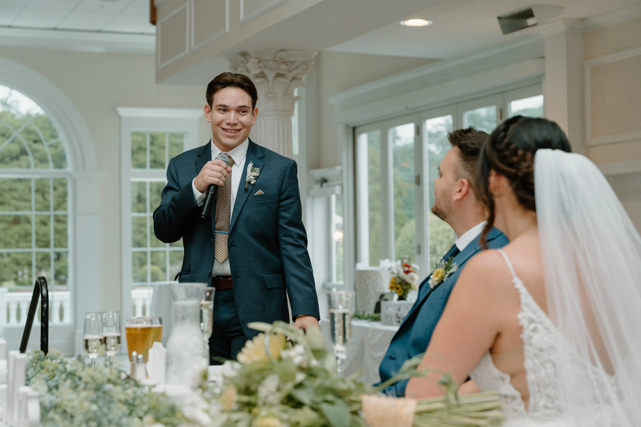 A young man giving a speech at a wedding reception, standing with a microphone, while the bride and groom are seated and listening during a wedding at the Aqua Turf Club in Connecticut. 