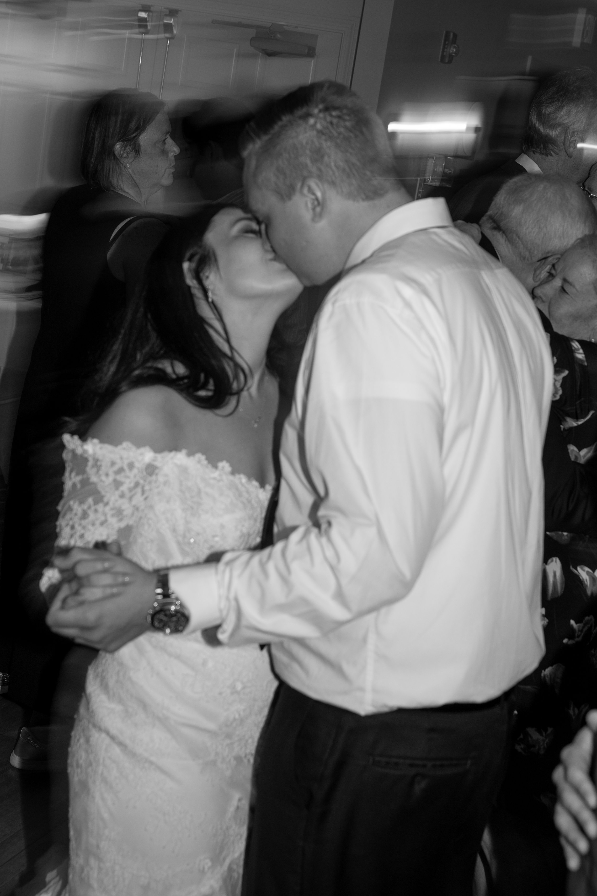 A bride and groom share a kiss during their wedding reception, surrounded by guests, in a black and white photo.