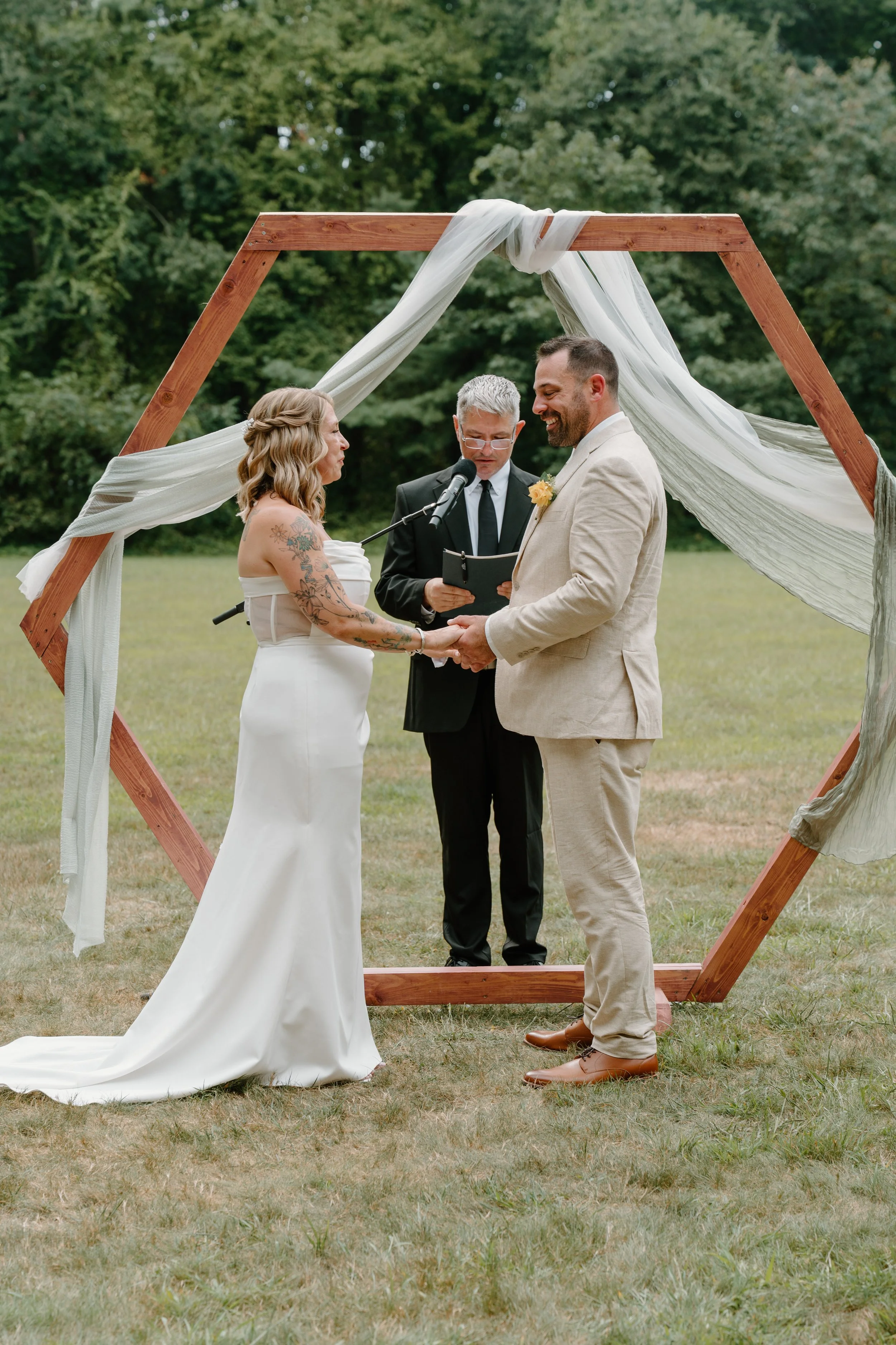 A couple getting married outdoors under a wooden hexagon wedding arch decorated with white fabric, with an officiant standing behind them for a wedding at Tunxis Country Club in Connecticut. 
